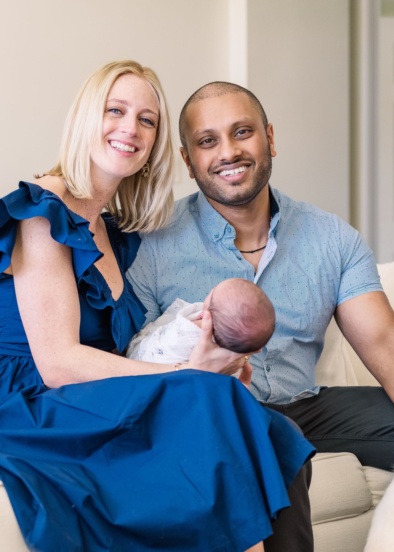 Couple in blue dress and button-down shirt holding their newborn baby on couch.