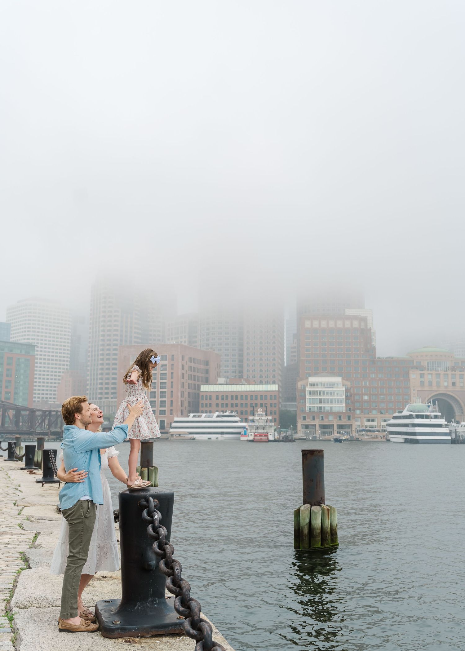 Family enjoying playful moments along a harbor chain fence with foggy Boston skyline.