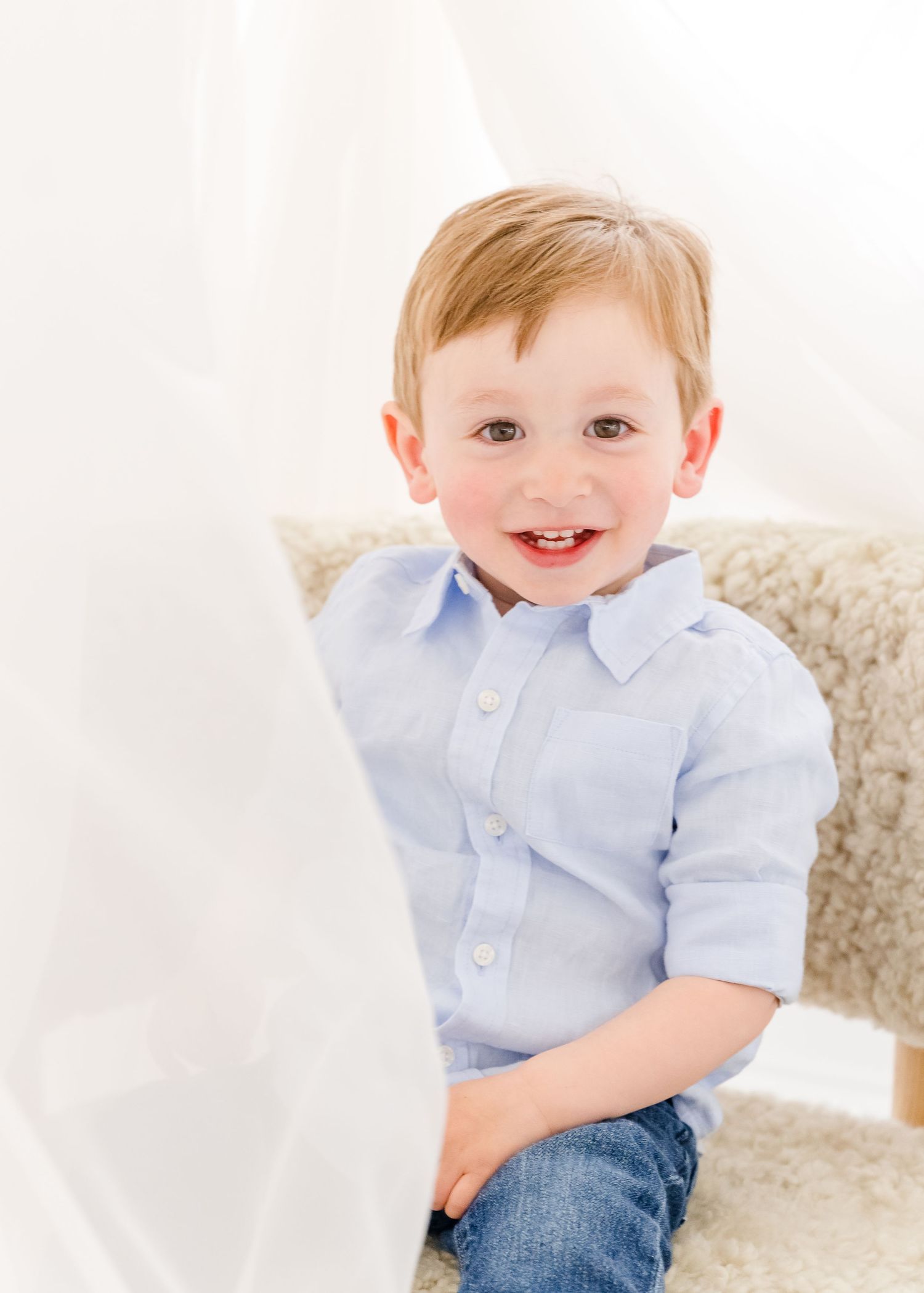 Small child wearing a light blue button-down shirt and jeans sits on a cream-colored chair with white background.
