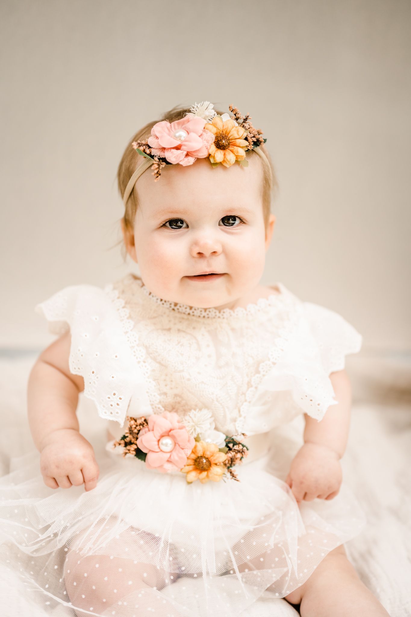 Baby in white lace dress and floral headband smiling at camera.