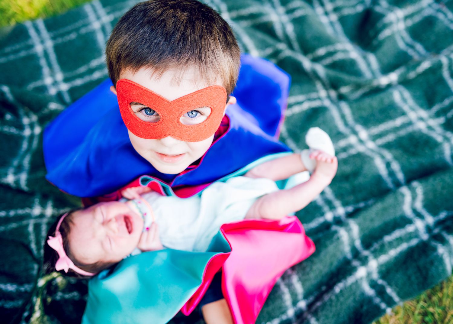 Child in superhero costume lying on plaid blanket outdoors.
