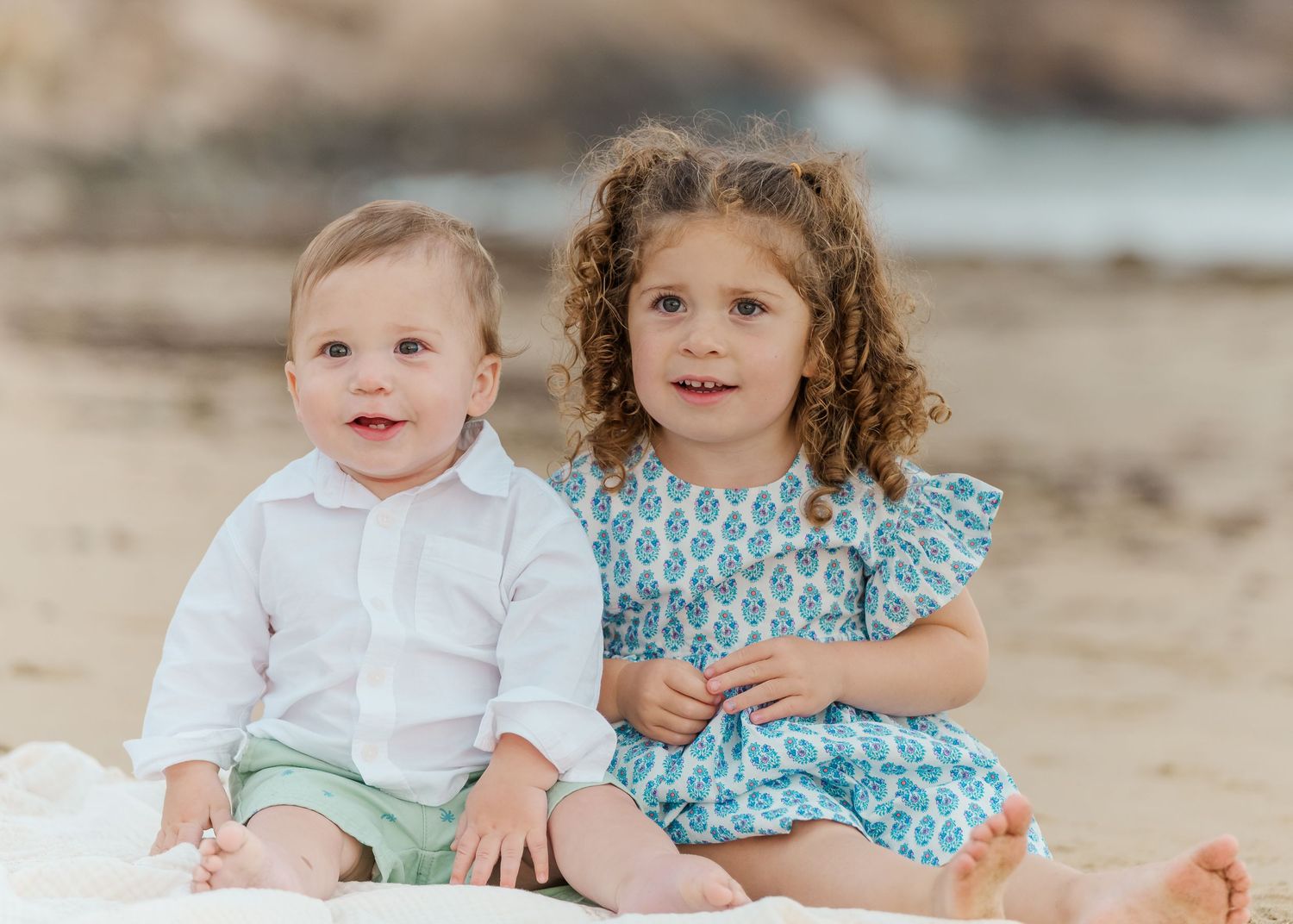 Two young children sit together on the beach wearing coordinated blue and white outfits.