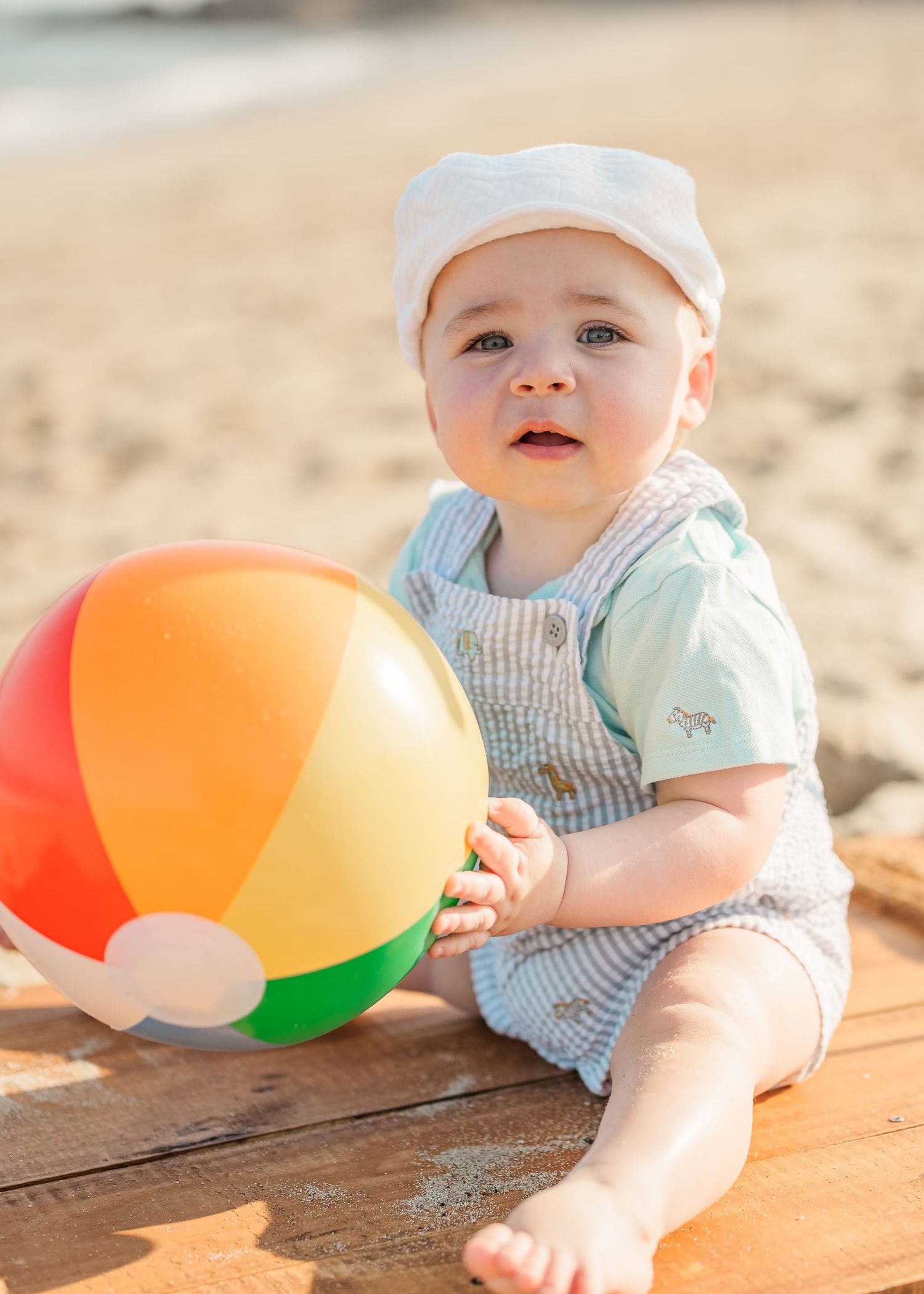 Baby in striped outfit playing with colorful beach ball on wooden deck in summer sunshine.