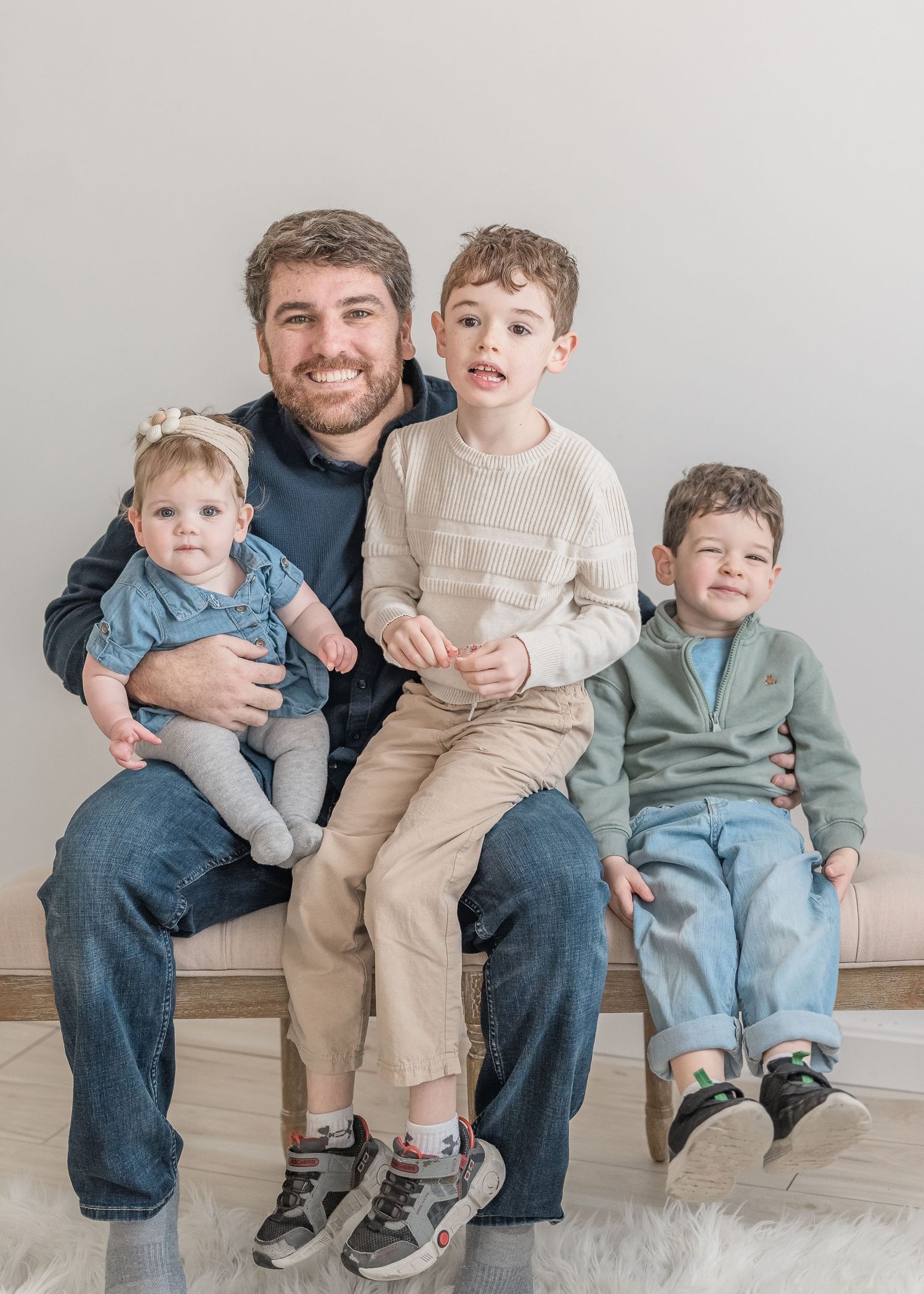 A family sitting on a bench against a white wall wearing coordinated outfits in blue and beige colors.