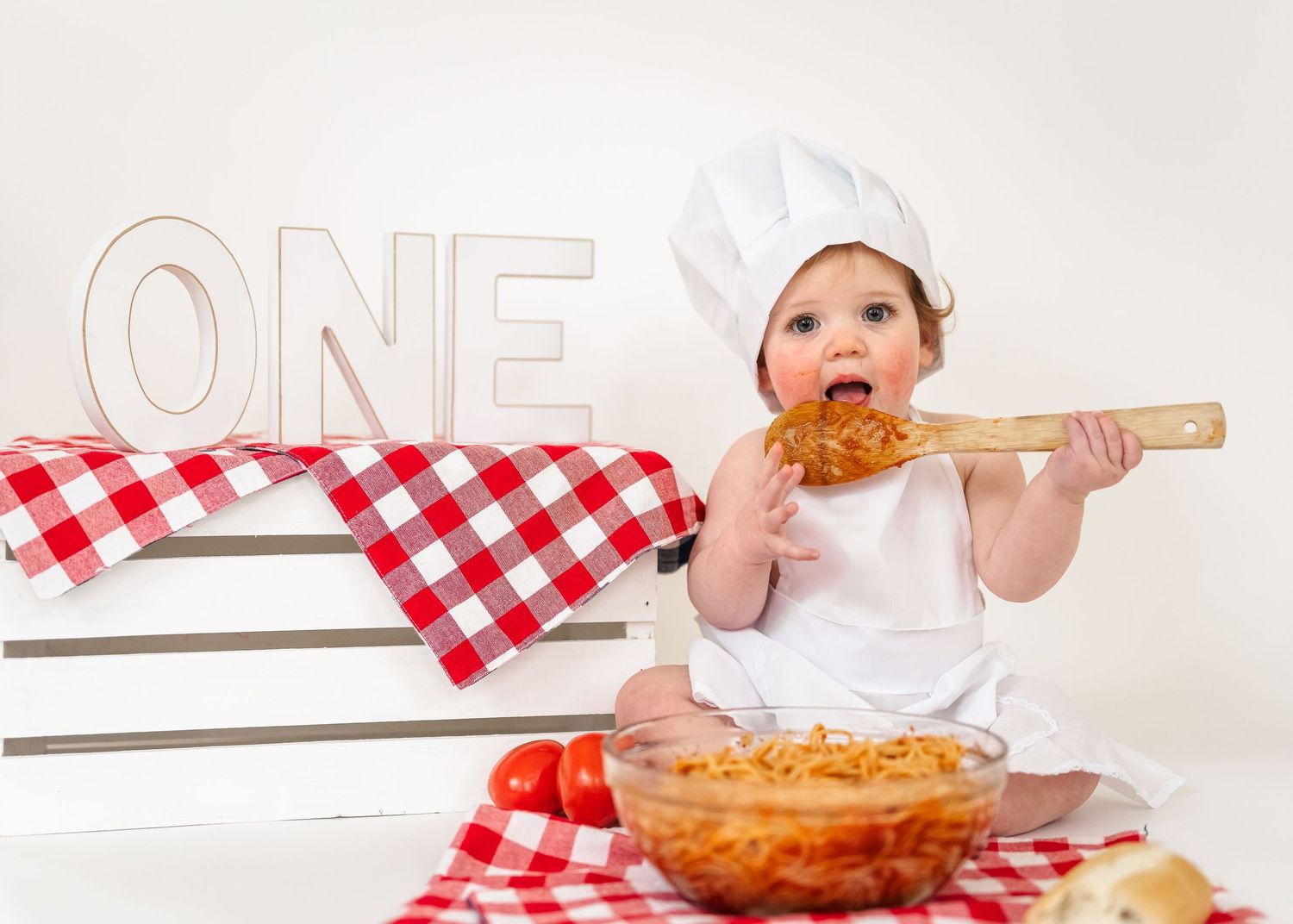 Spaghetti Smash first birthday. A baby chef in white uniform eats spaghetti from a wooden spoon on a red checkered cloth.
