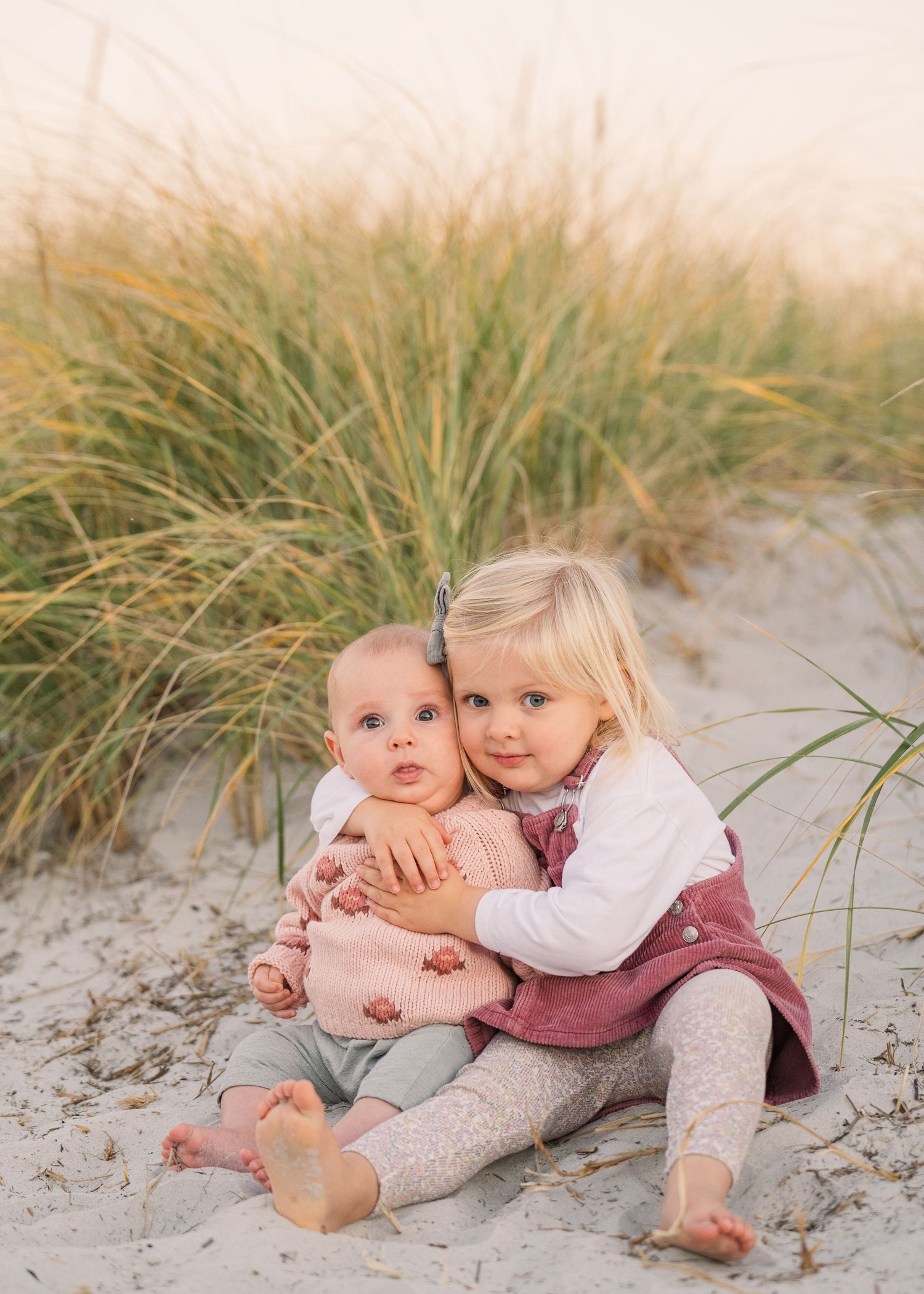 Two young children in matching outfits sit together on sandy beach with coastal grass in background.