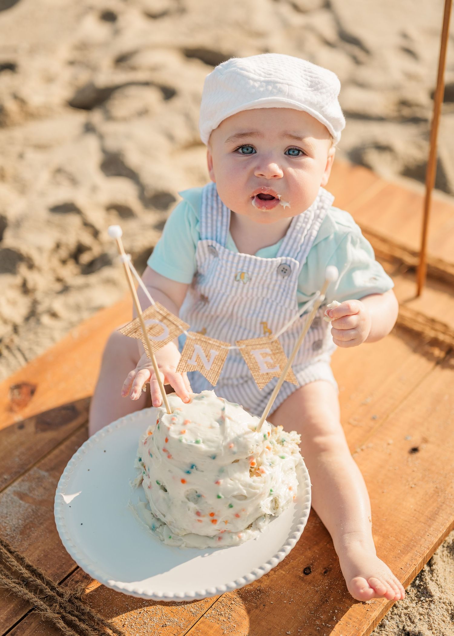 Baby wearing blue and white striped outfit smiles while playing with smash cake on wooden surface at beach.