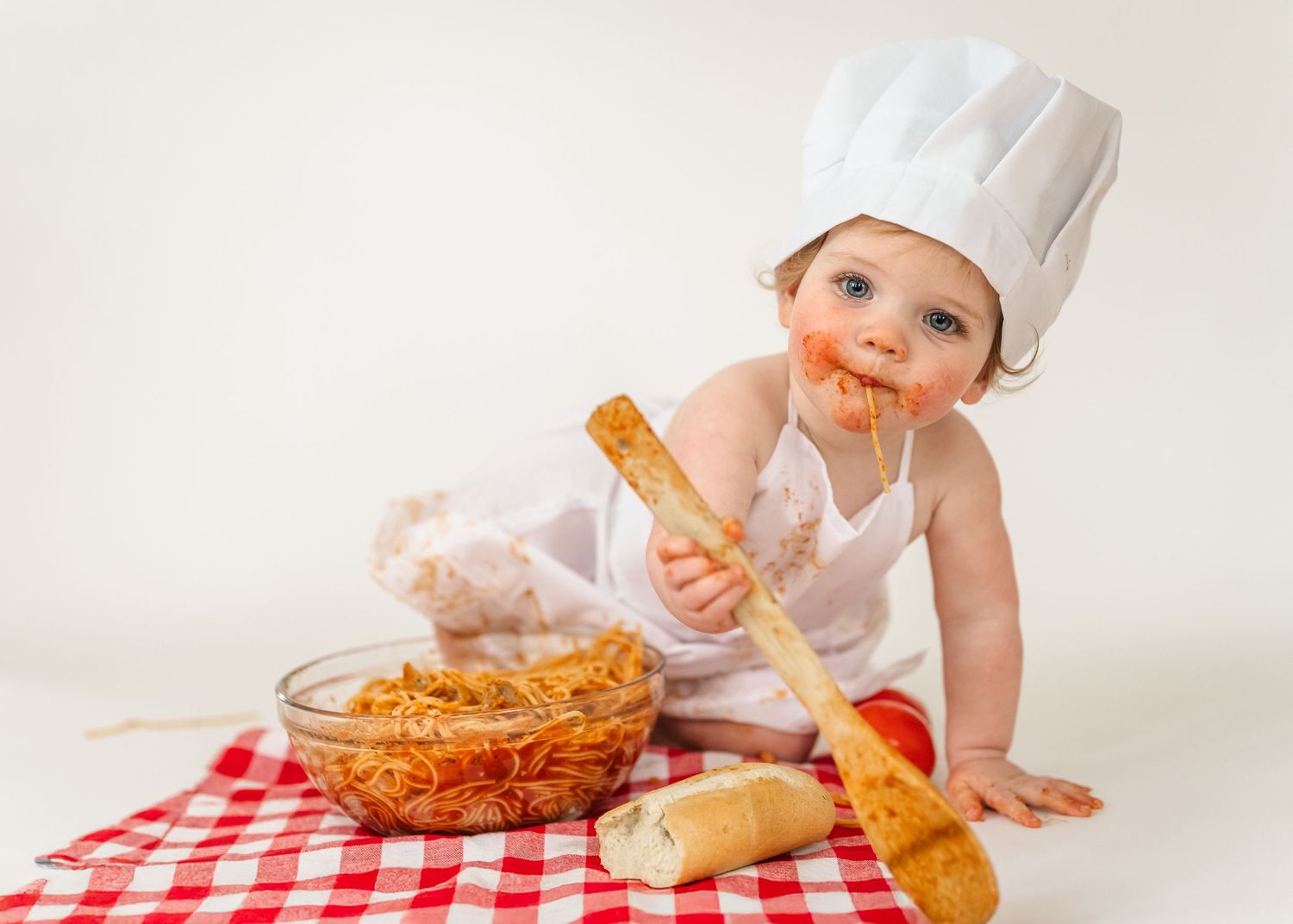 Spaghetti Smash first birthday. Small chef in white hat and apron plays with spaghetti and bread on checkered tablecloth.