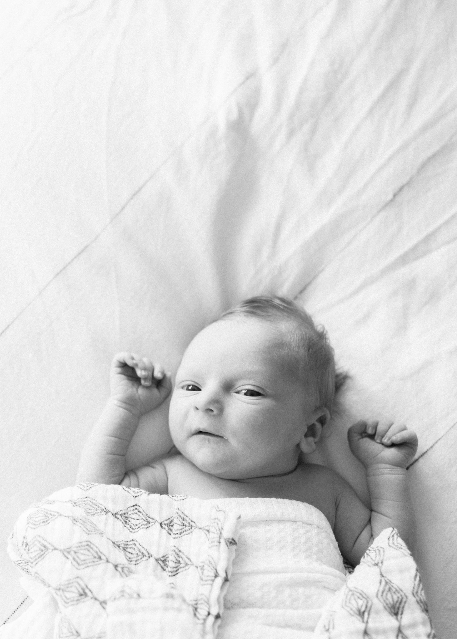 Black and white portrait of baby lying on white bedding with arms raised.
