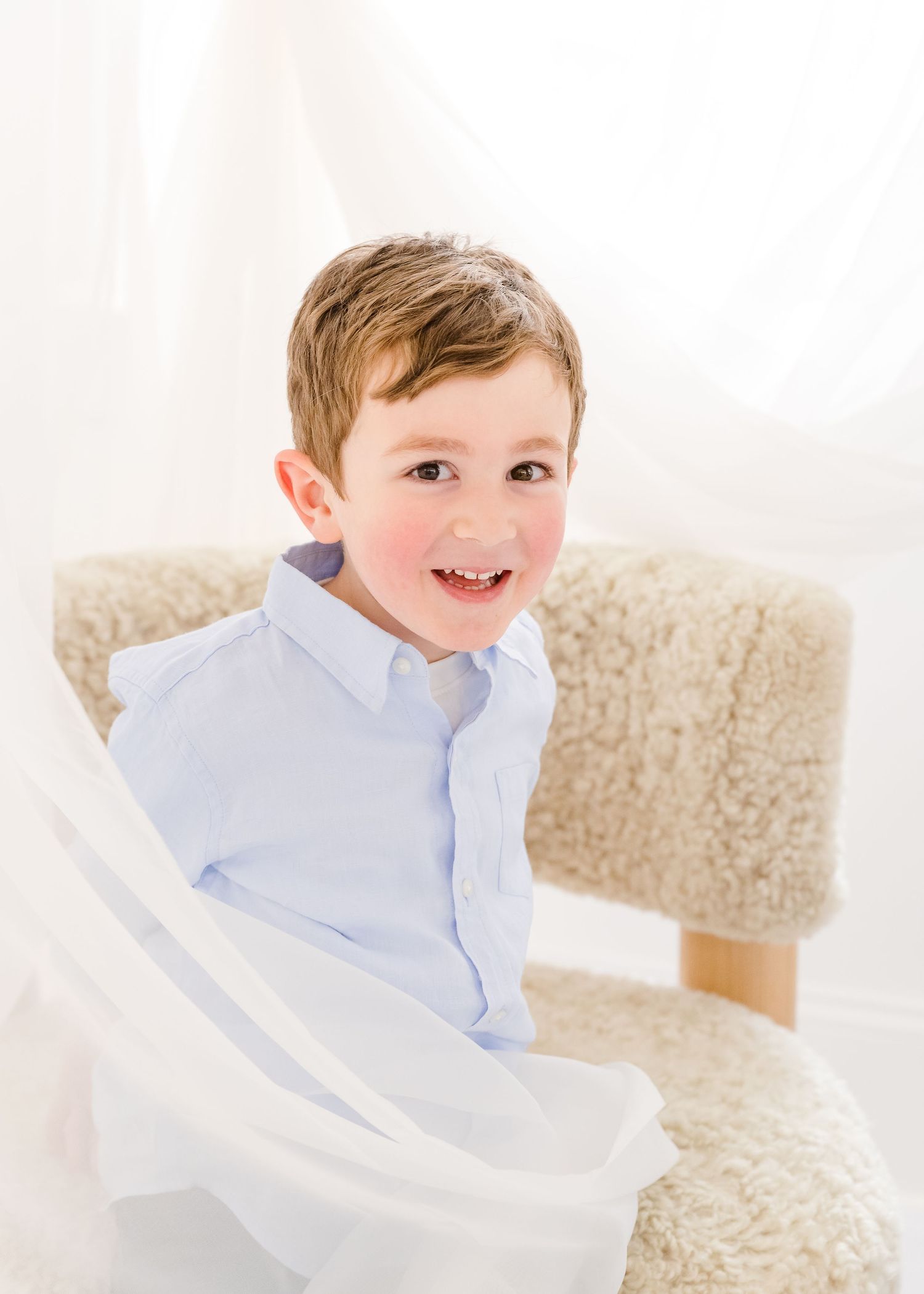 A young child in a light blue button-down shirt smiles while sitting in a cozy cream-colored chair.