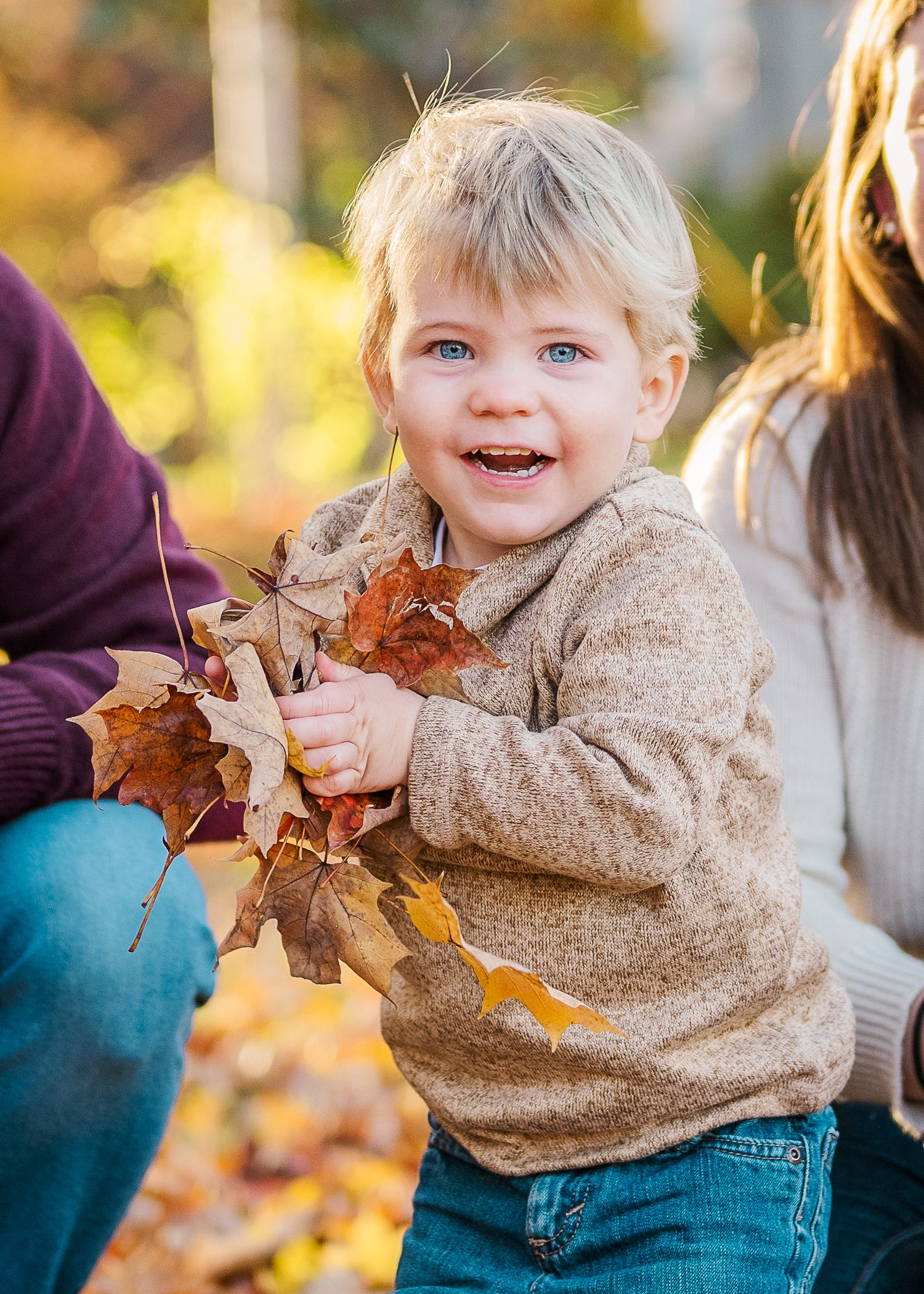 A toddler in a beige sweater laughs while playing with fallen autumn leaves.