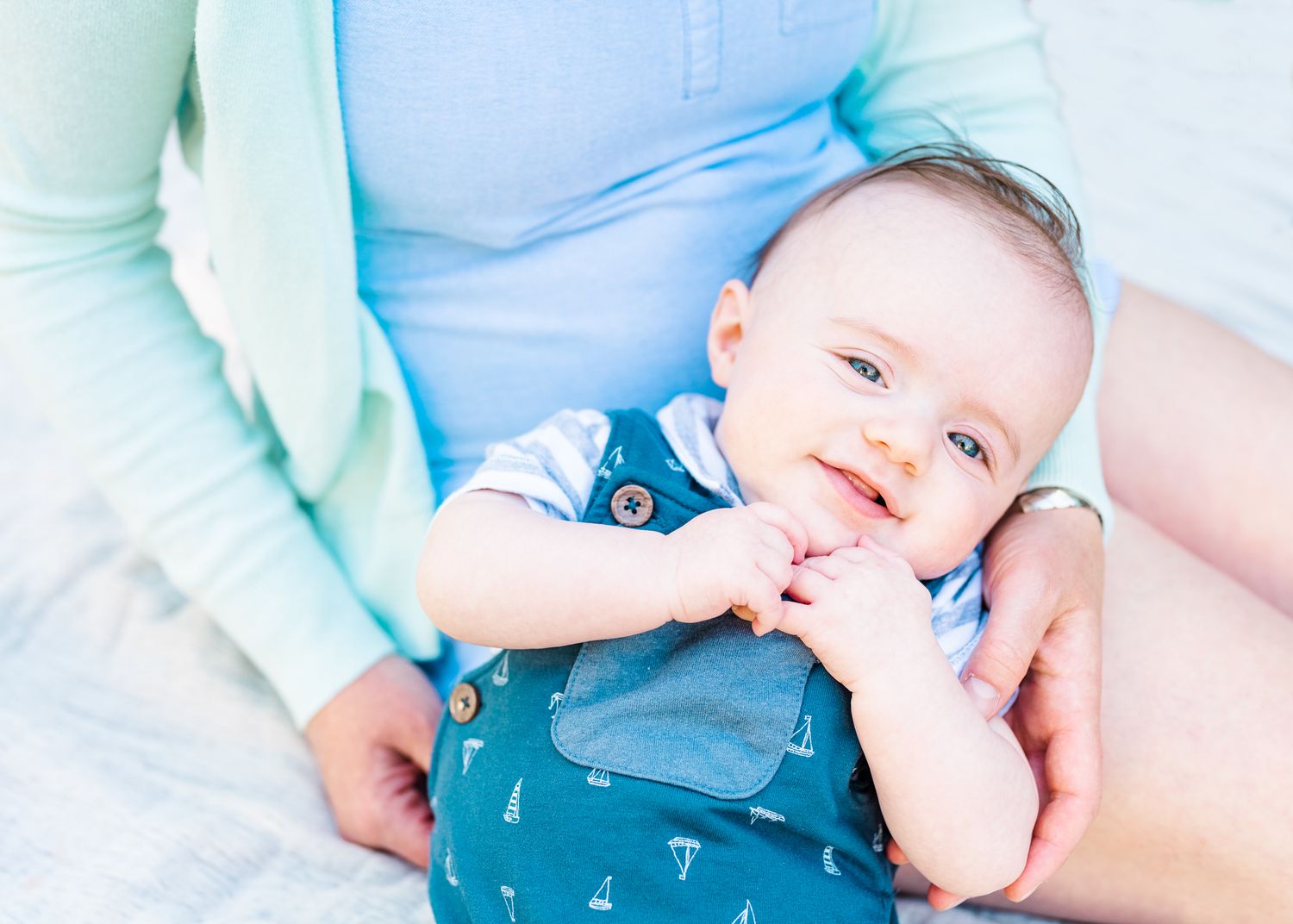 Baby wearing blue outfit smiles while being held against light blue background.
