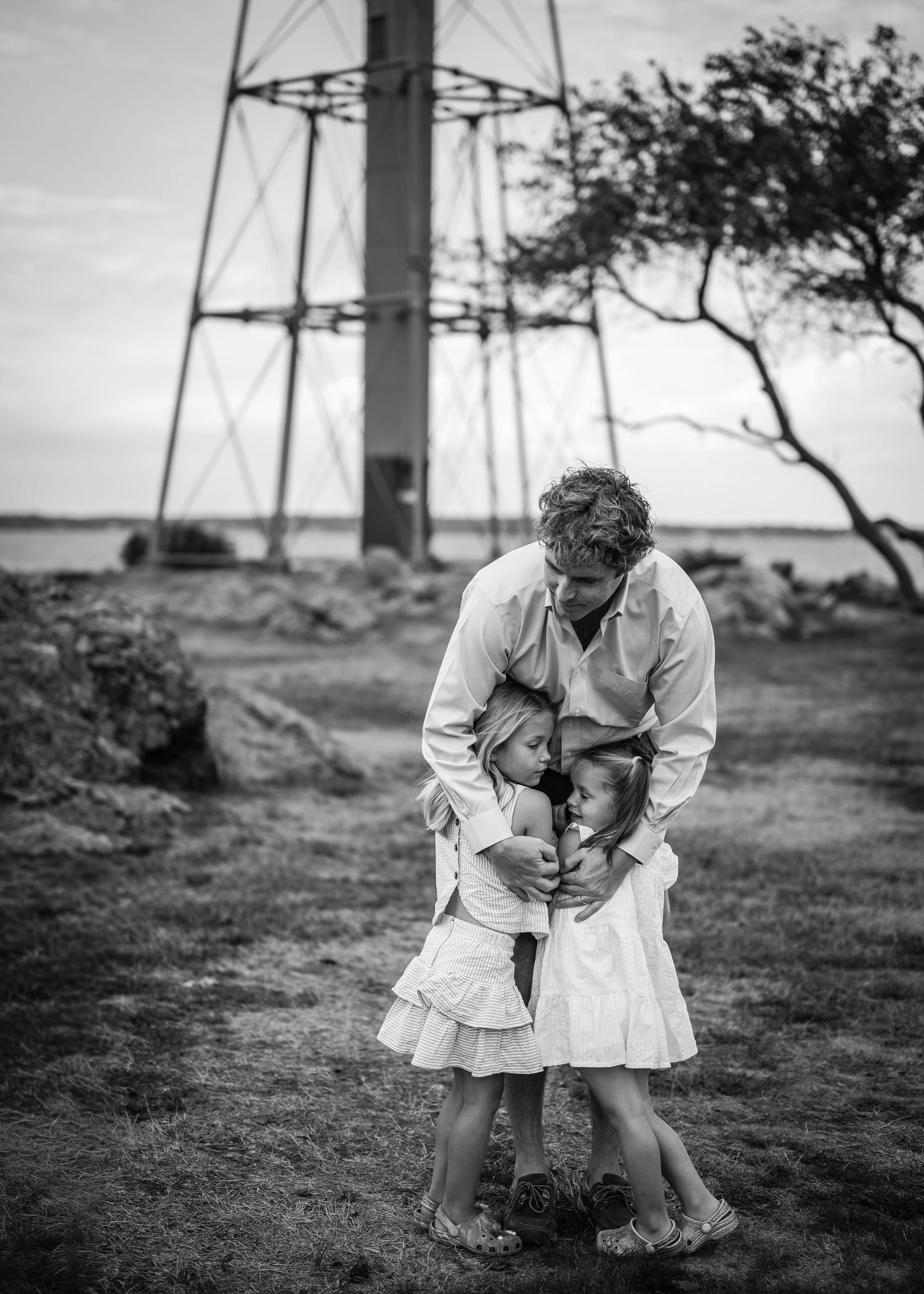 A tender black and white moment captured near a lighthouse with two people embracing on rocky ground.