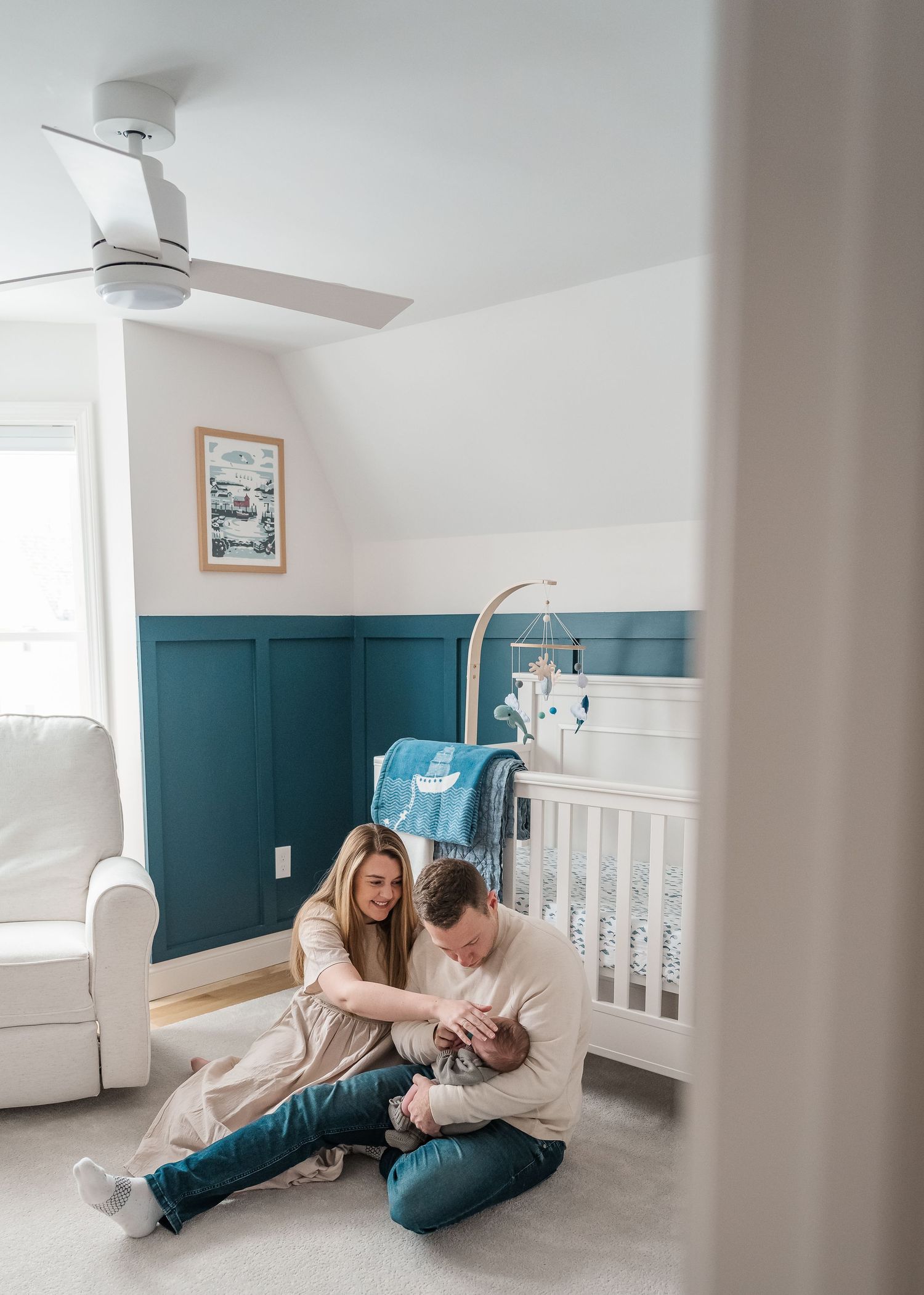 Nursery room with teal wainscoting features parents sitting on floor with baby near white crib.