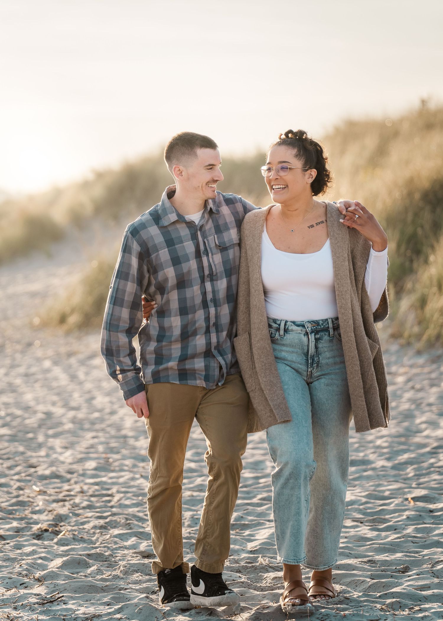 A couple walks together on a beach at sunset, wearing casual outfits and sharing a happy moment.