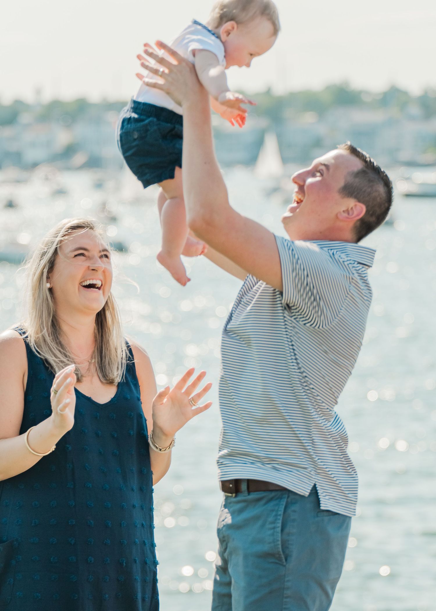 A couple on the beach laughs joyfully while lifting a baby in a striped shirt and blue shorts up in the air.