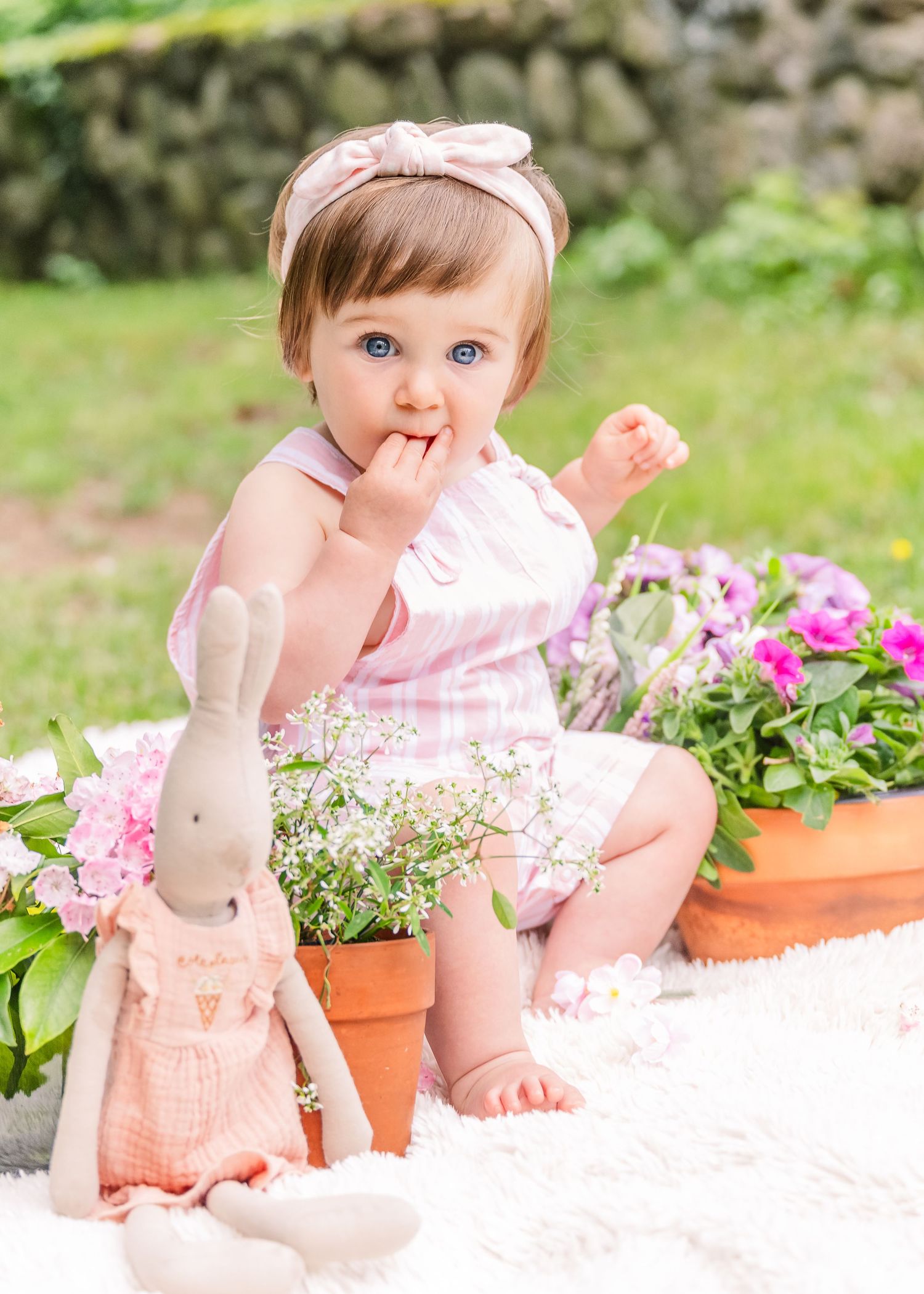 Child in pink dress sitting by flower pots with stuffed bunny toy outdoors.