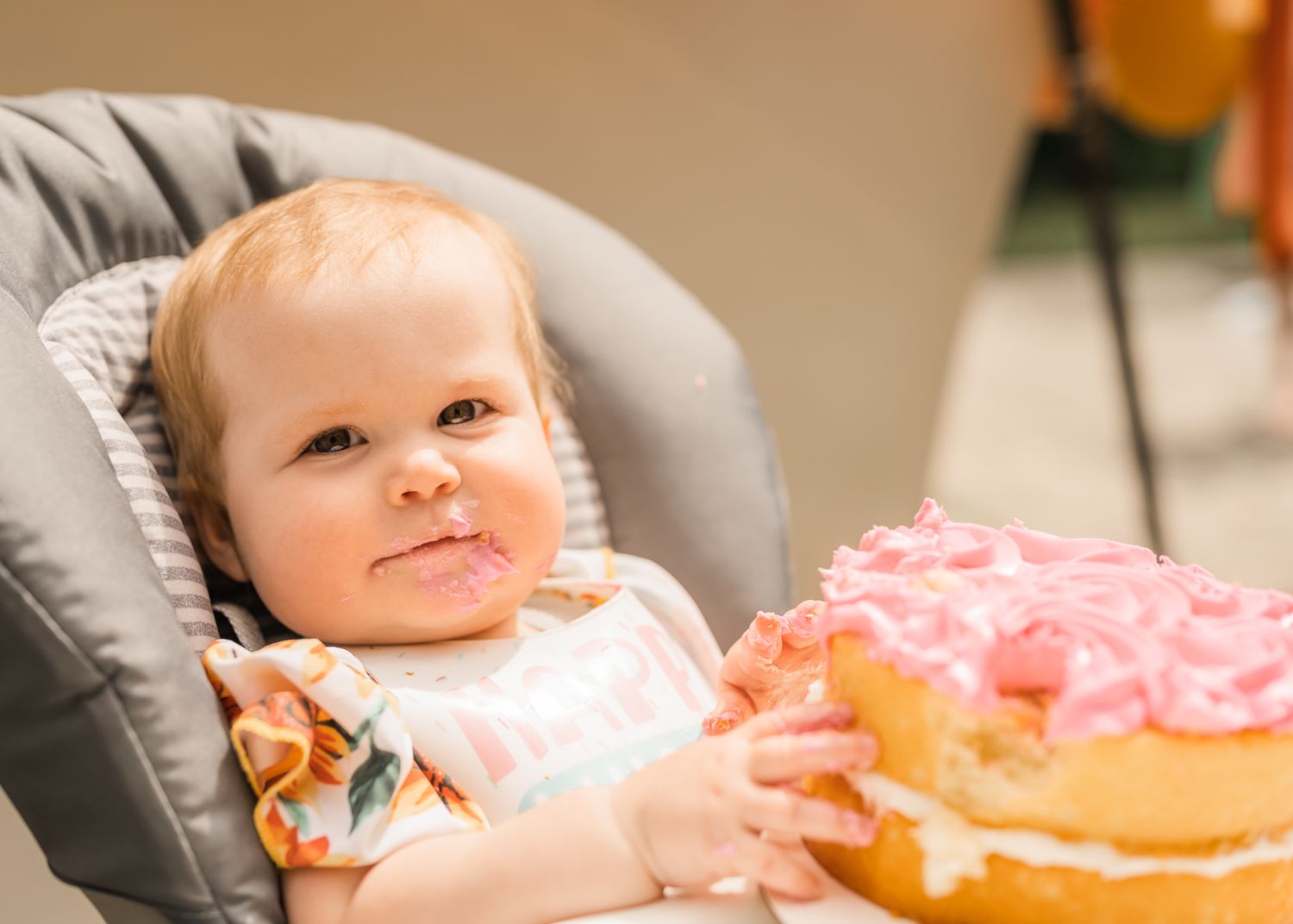 Baby in high chair admiring pink frosted birthday cake at celebration.