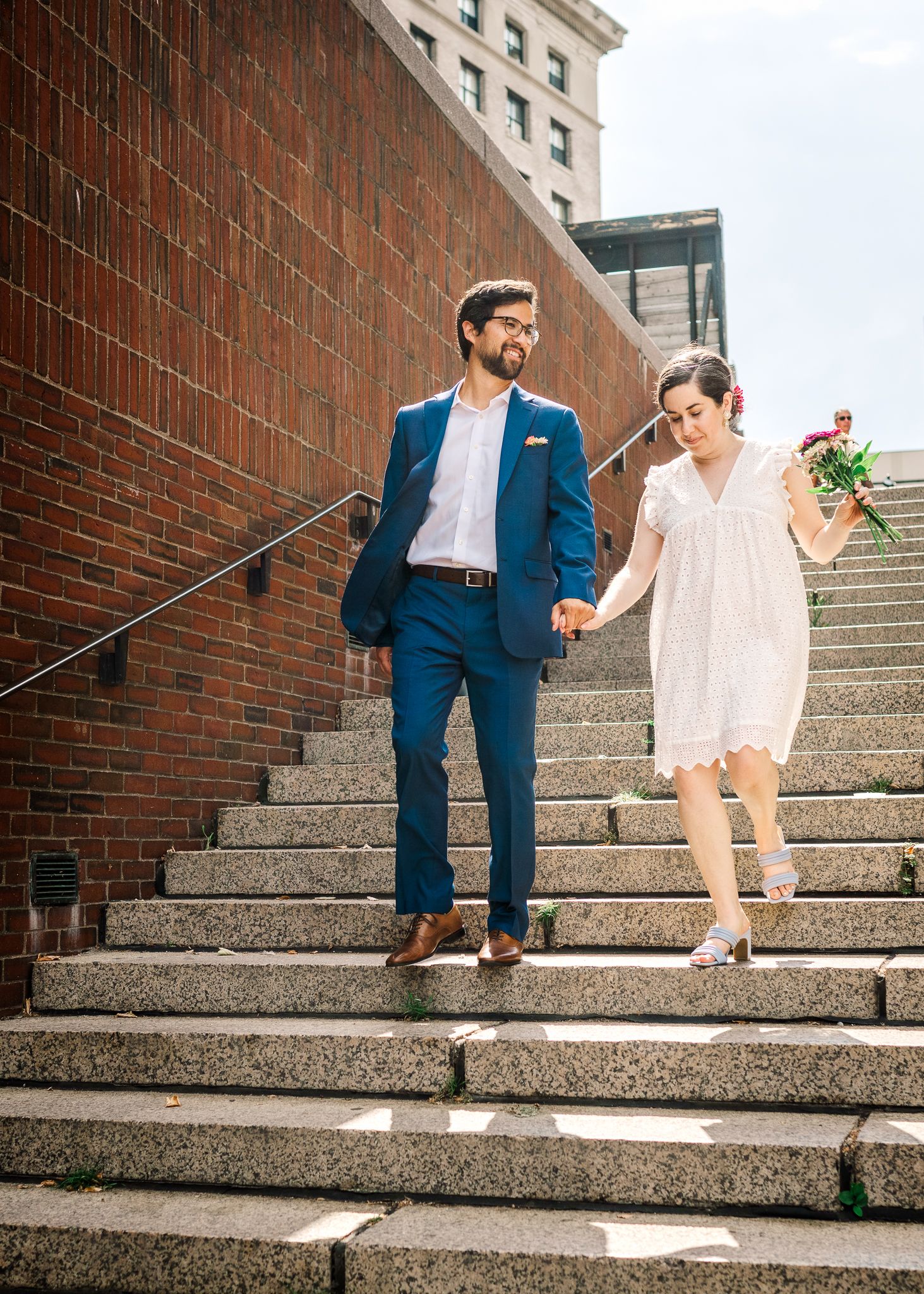 Couple descending concrete stairs together, dressed elegantly in blue suit and blush dress.