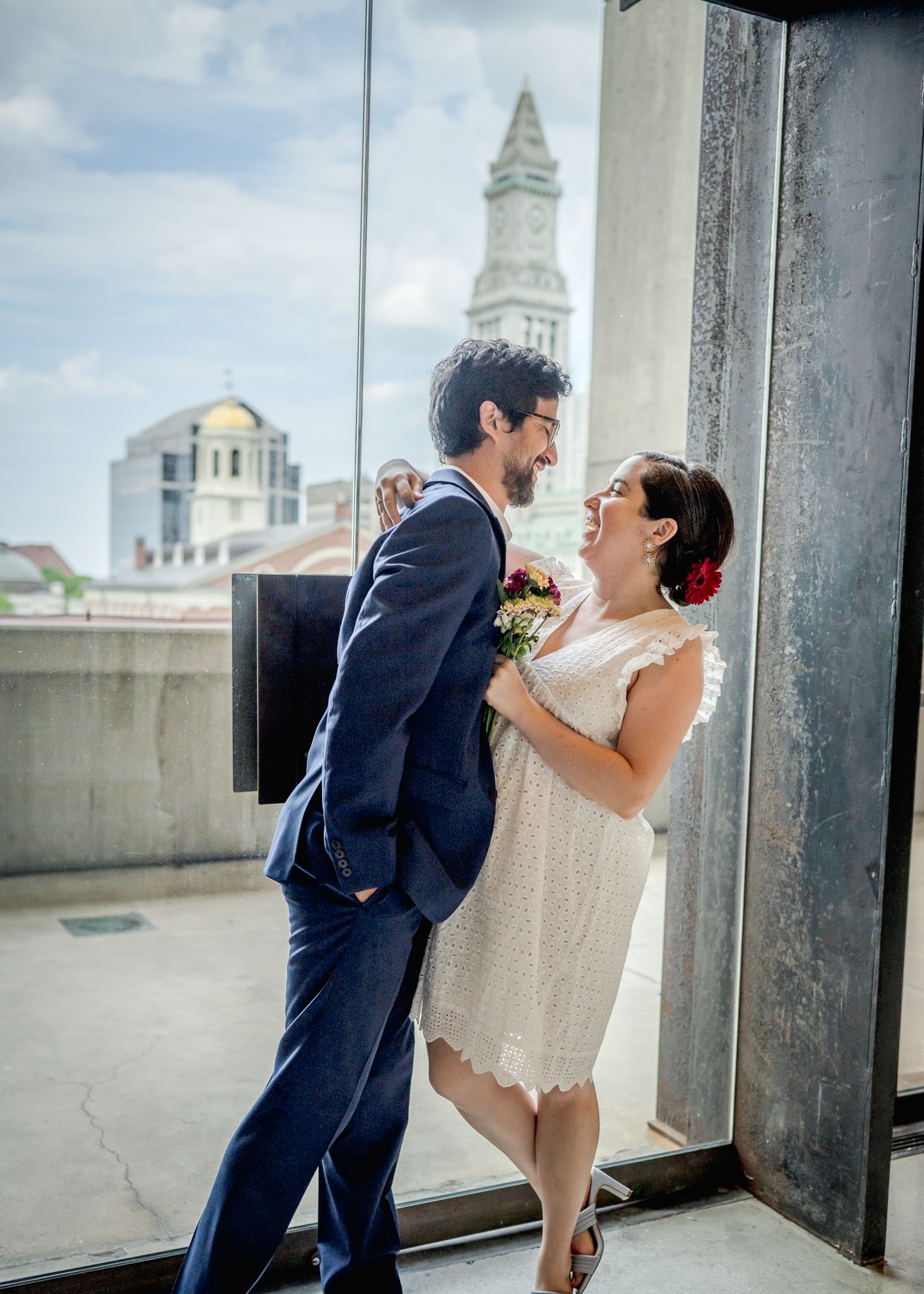 Wedding couple poses playfully by window with Boston cityscape in background.