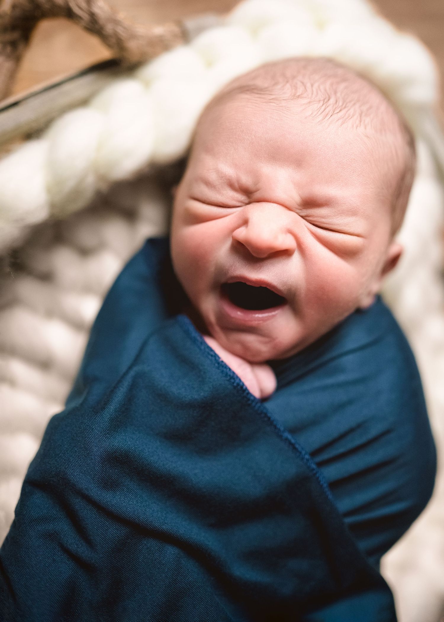 A swaddled newborn baby with scrunched up expression lets out a big yawn.