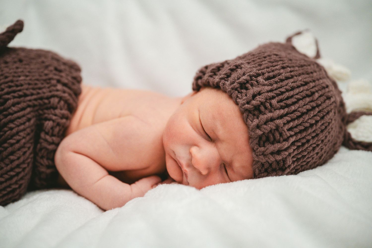 A sleeping newborn wearing a brown knit hat rests peacefully on white bedding.