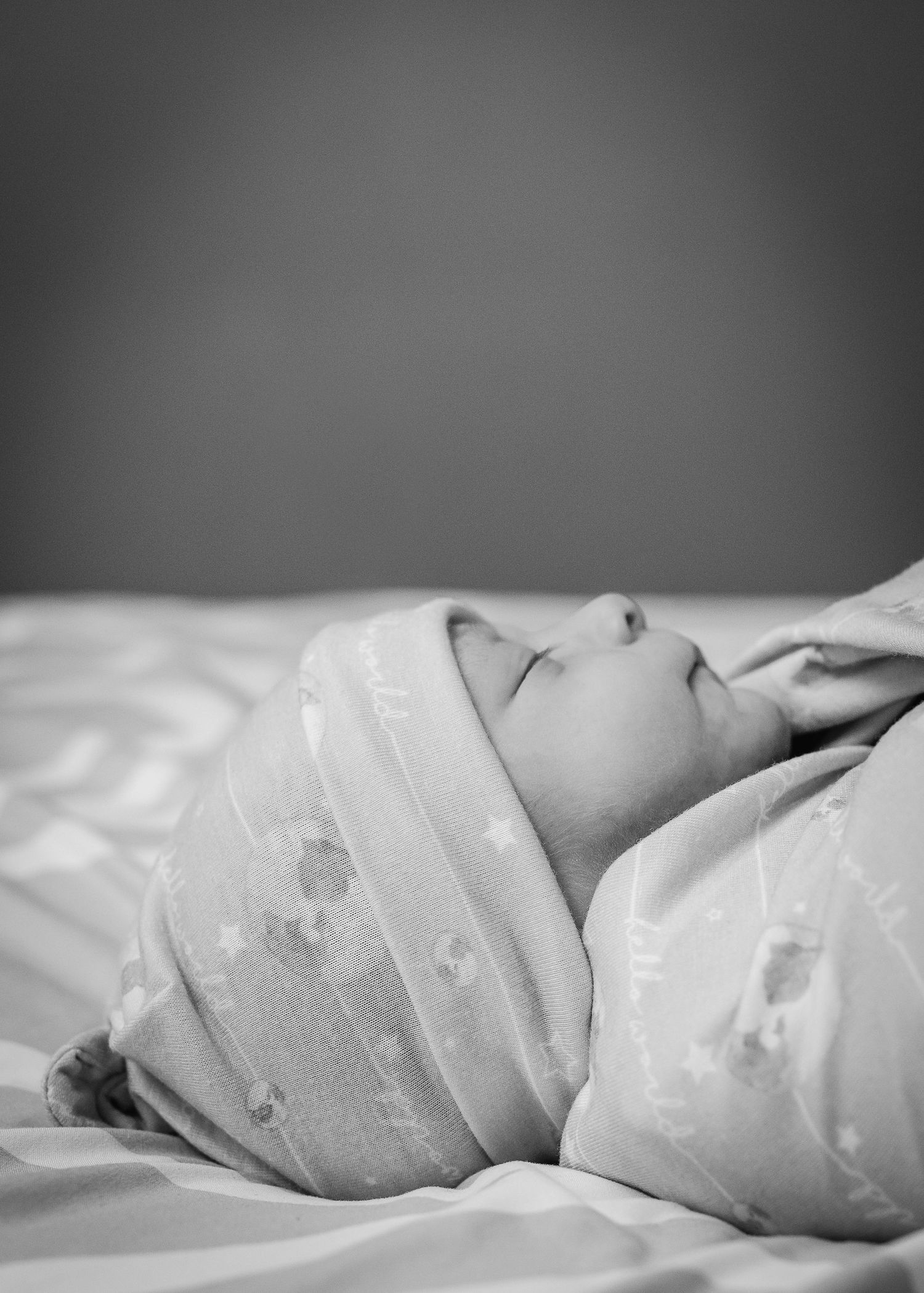 A sleeping newborn wearing delicate white fabric rests peacefully on white linens in black and white.