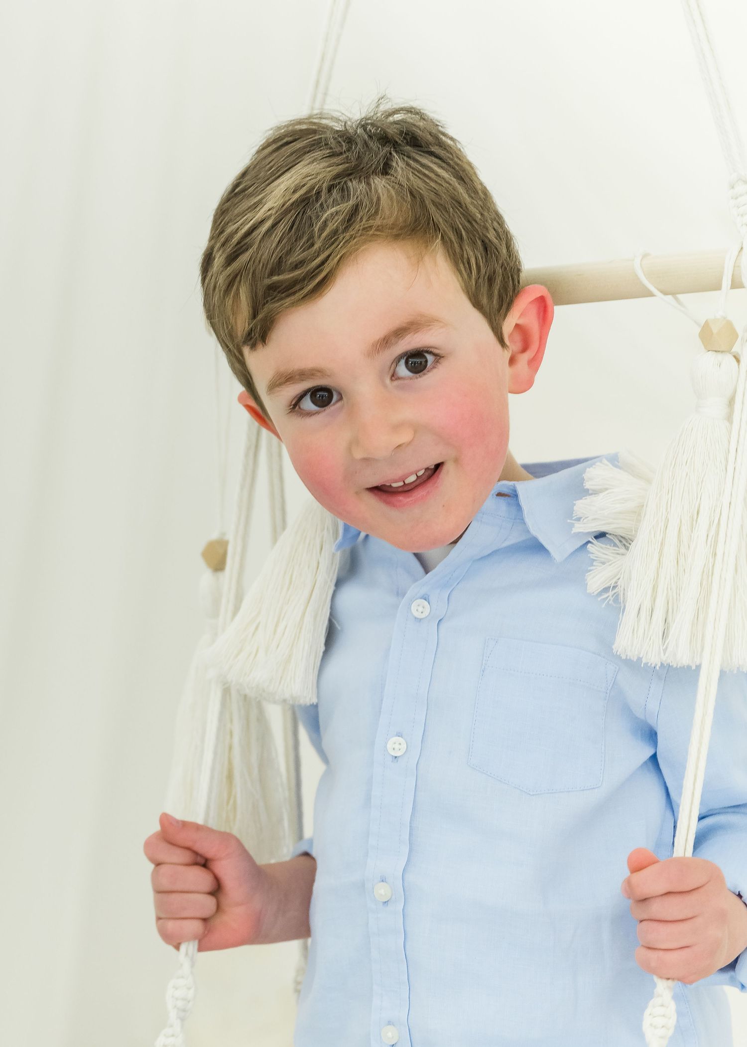Young child wearing a light blue button-down shirt smiles while standing next to white curtains or drapes.