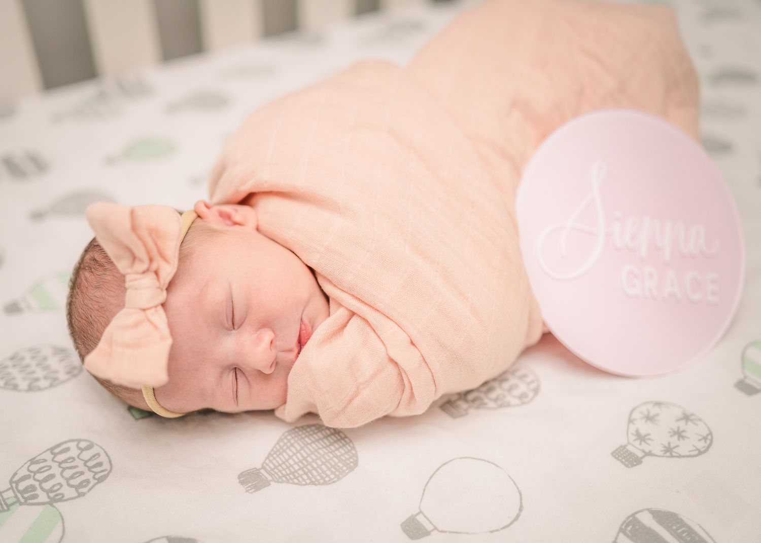 Sleeping newborn in peach wrap on patterned crib sheet with pink circle decoration.