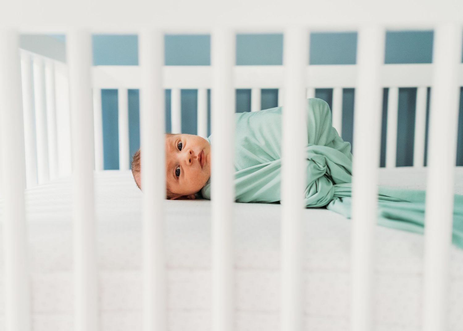 Baby in mint green clothes lying in white wooden crib.