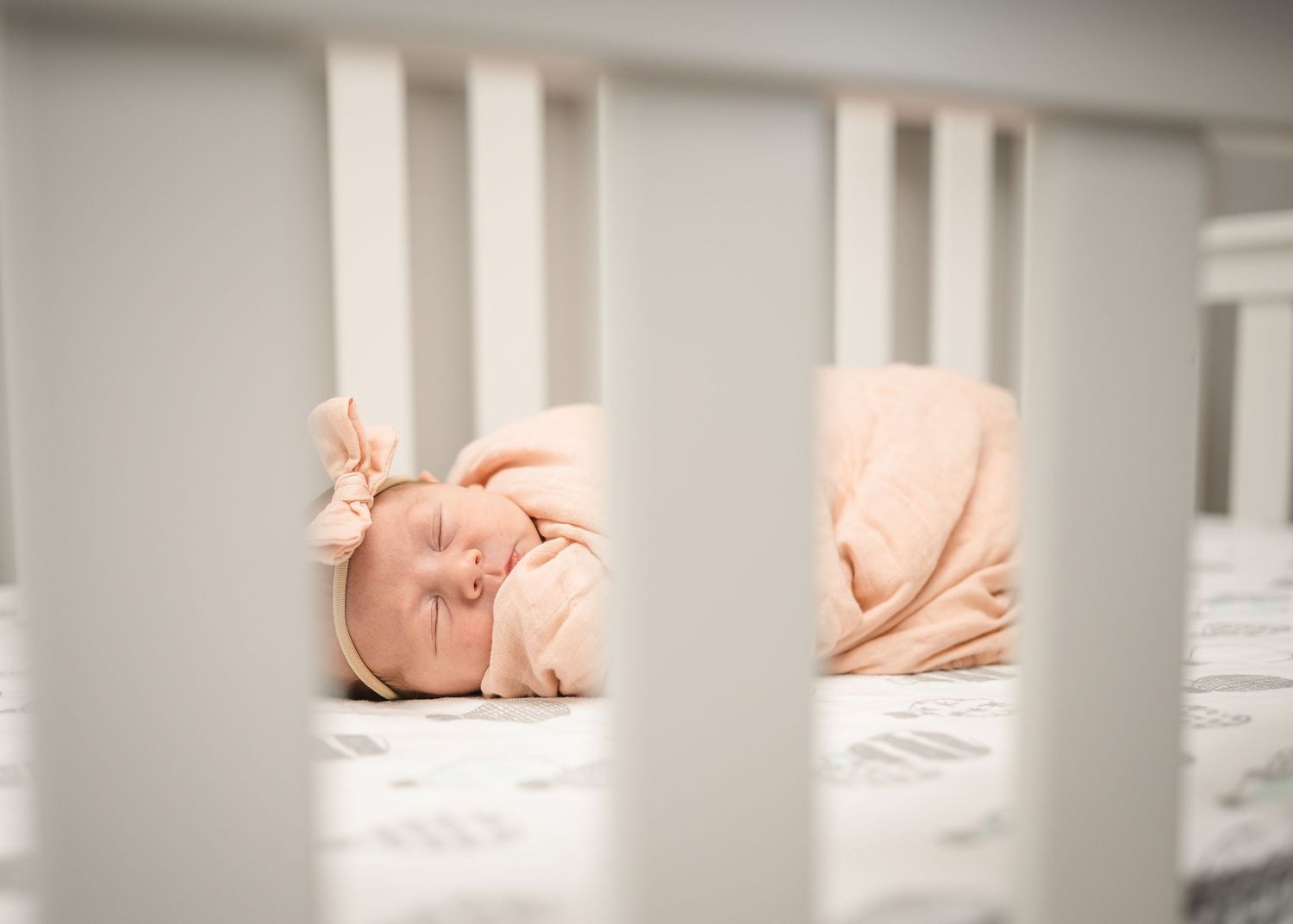 A sleeping baby in peach clothing viewed through white crib slats.