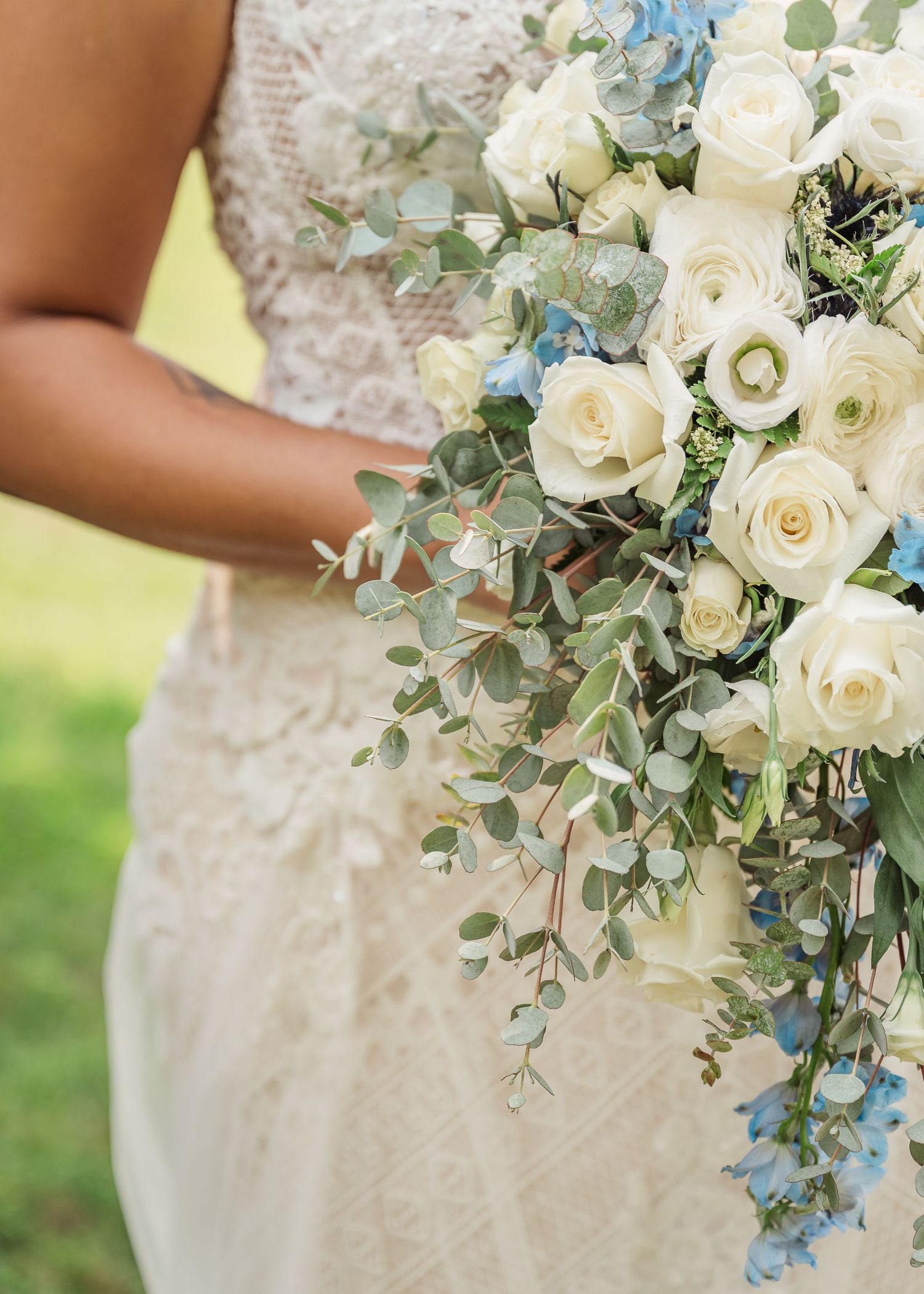 Elegant white and blue wedding bouquet with roses, delphinium, and eucalyptus against a lace wedding dress.