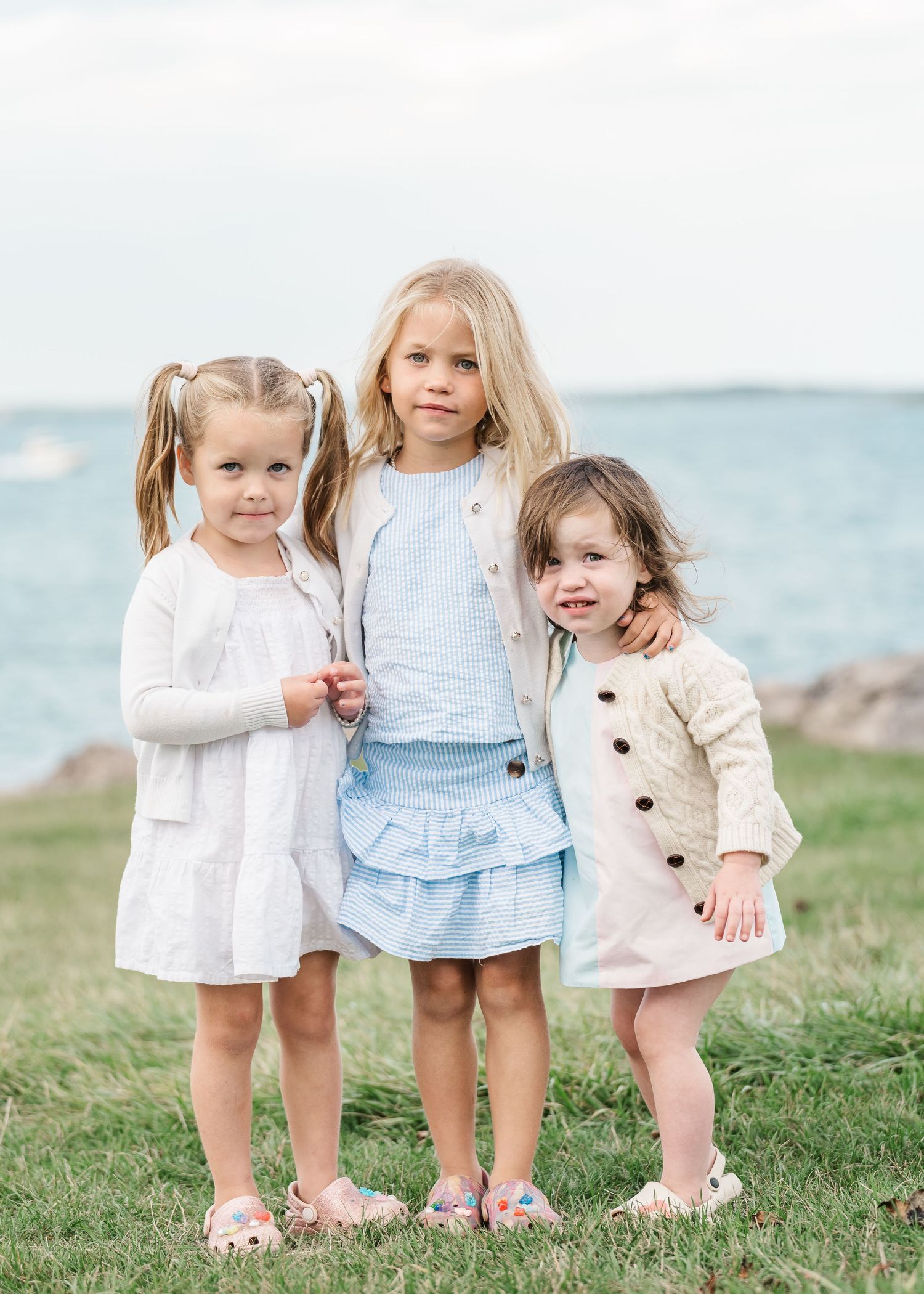 Three children in light colored dresses stand together on grassy hill overlooking water in matching outfits.