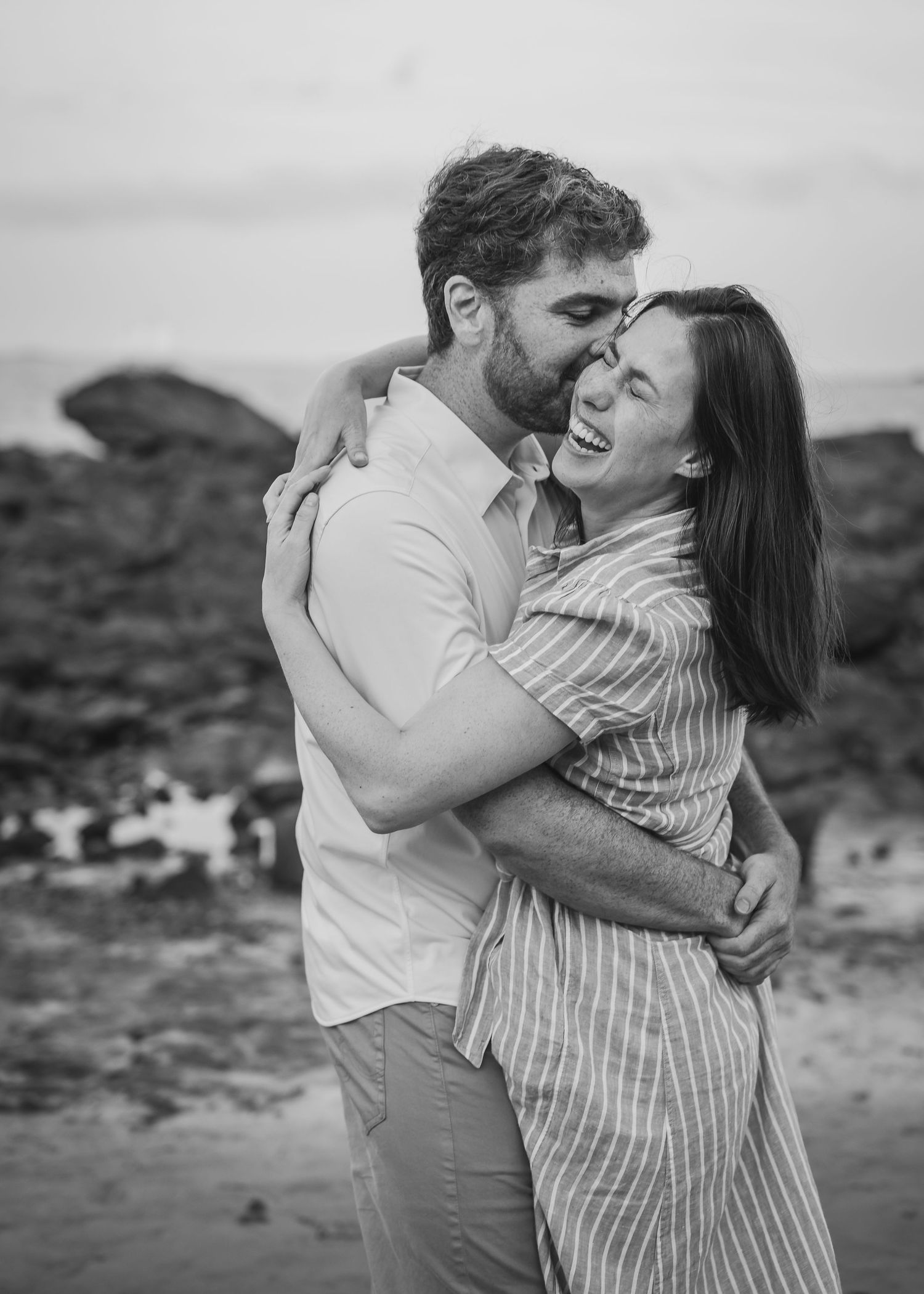 A candid black and white photo of a couple laughing and embracing on a rocky beach.