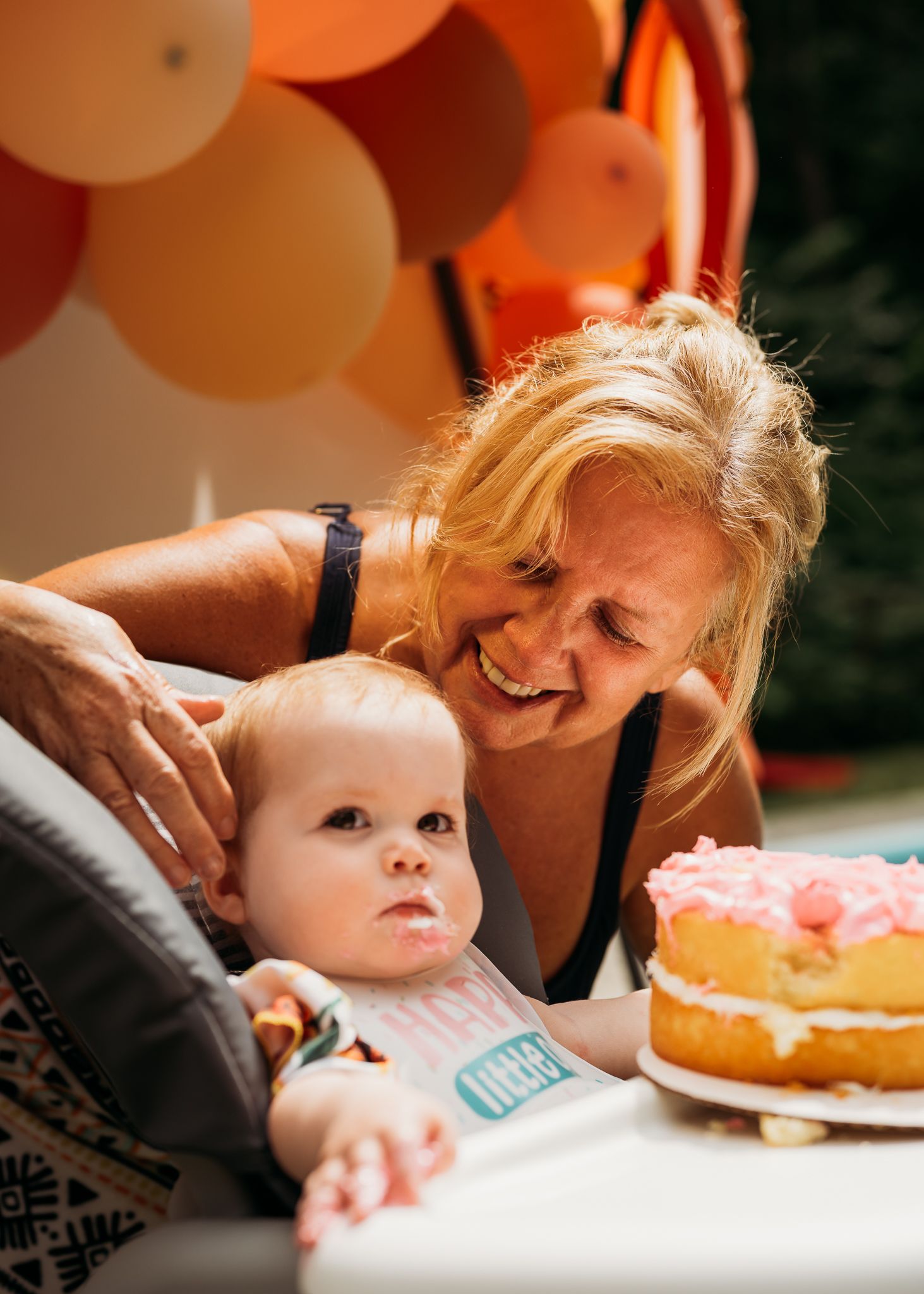Joyful celebration moment with baby in high chair near pink birthday cake and peach balloons.