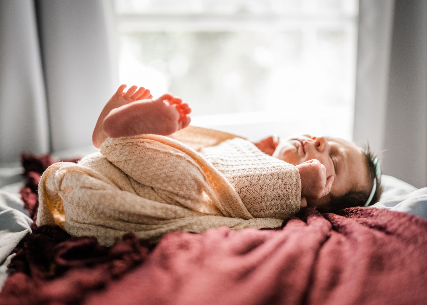 Newborn baby sleeping peacefully in white swaddle on burgundy blanket by window.