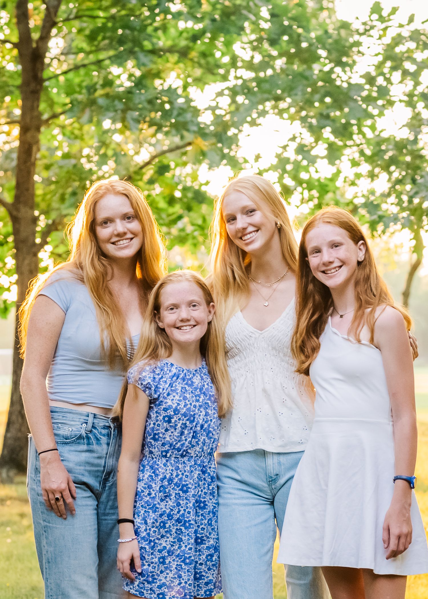 Four redheaded siblings stand together outdoors in casual summer attire against a leafy background.