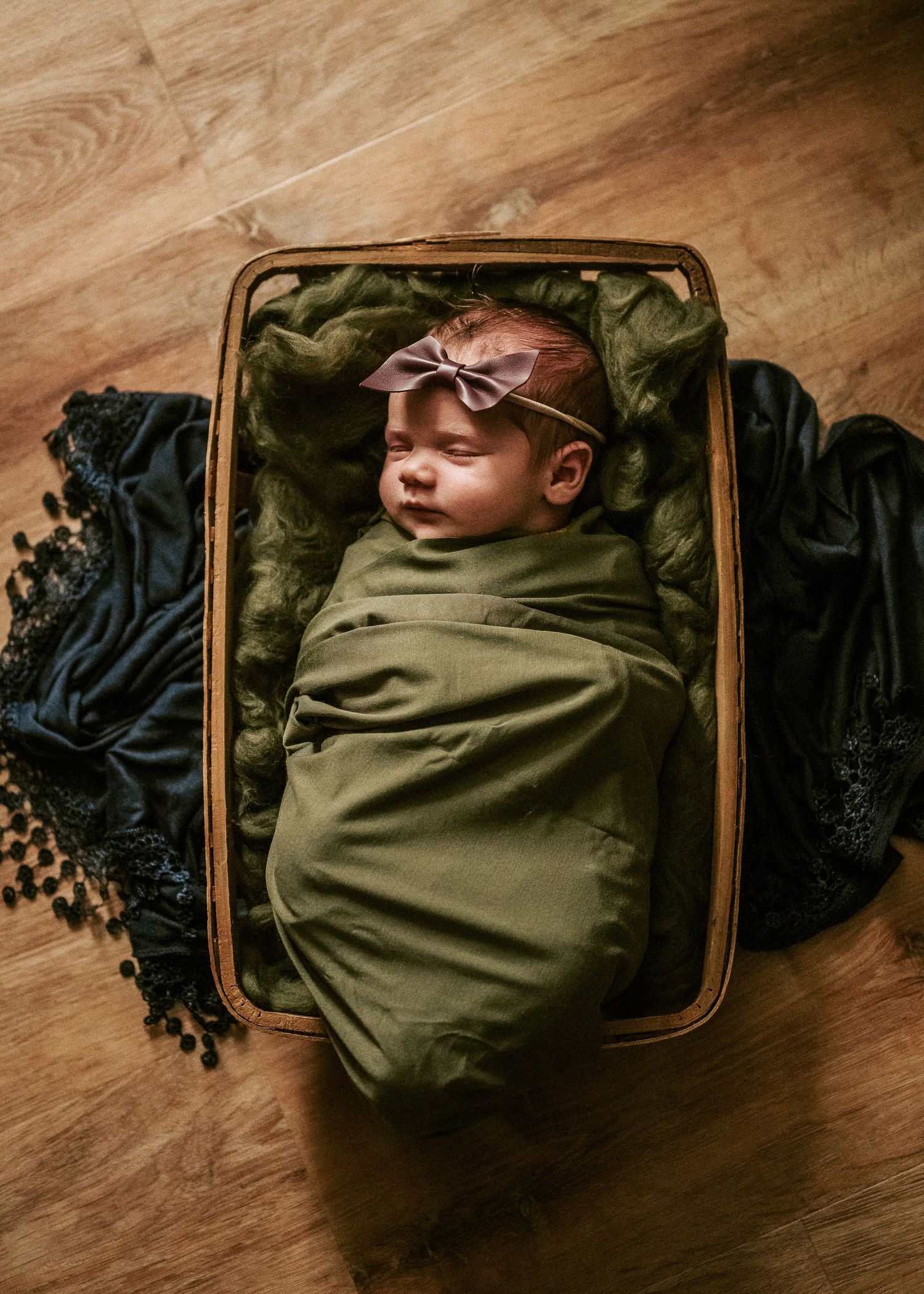 Sleeping newborn wrapped in olive green swaddle blanket rests peacefully in vintage brown leather suitcase on hardwood floor.