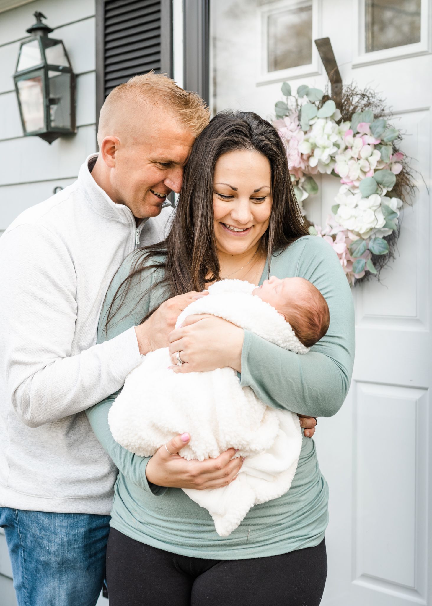 A couple shares joyful moment with their newborn baby outside their home with decorative wreath.