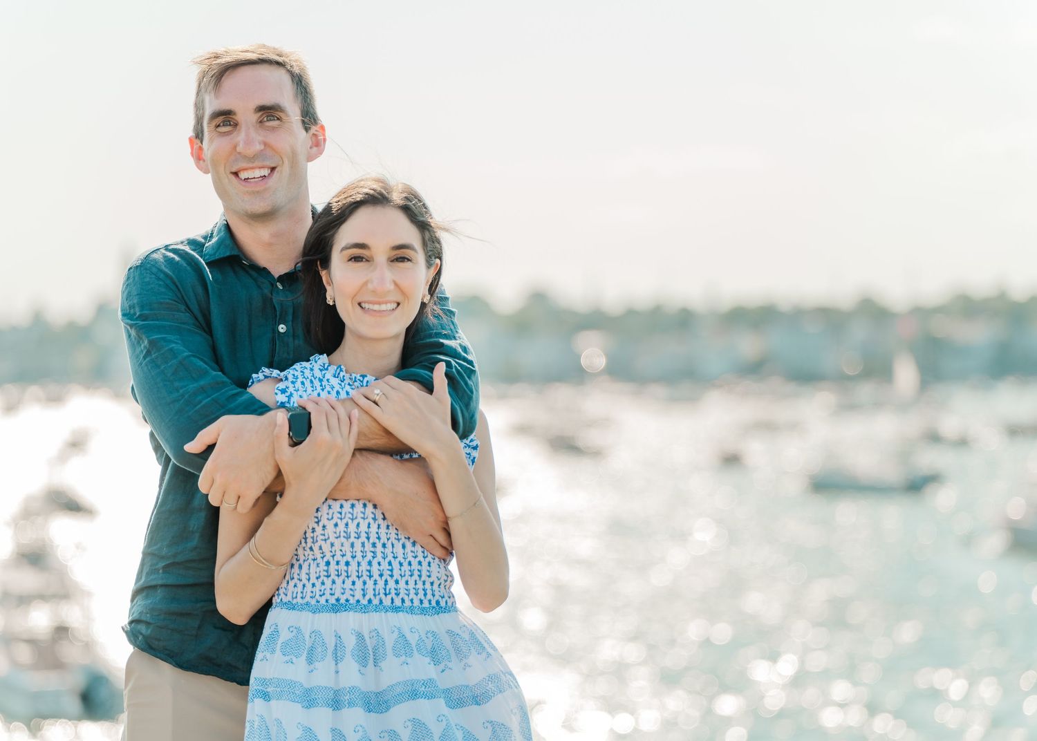 A couple embraces while standing on a beach with boats and sparkling water visible in the distance.