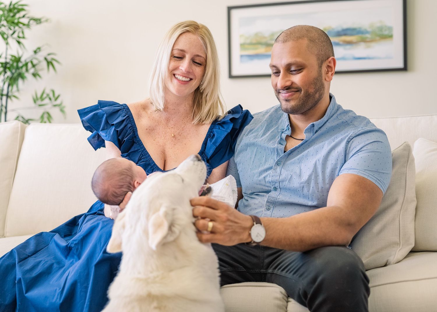 Family on couch with white dog watching as they hold their newborn baby.