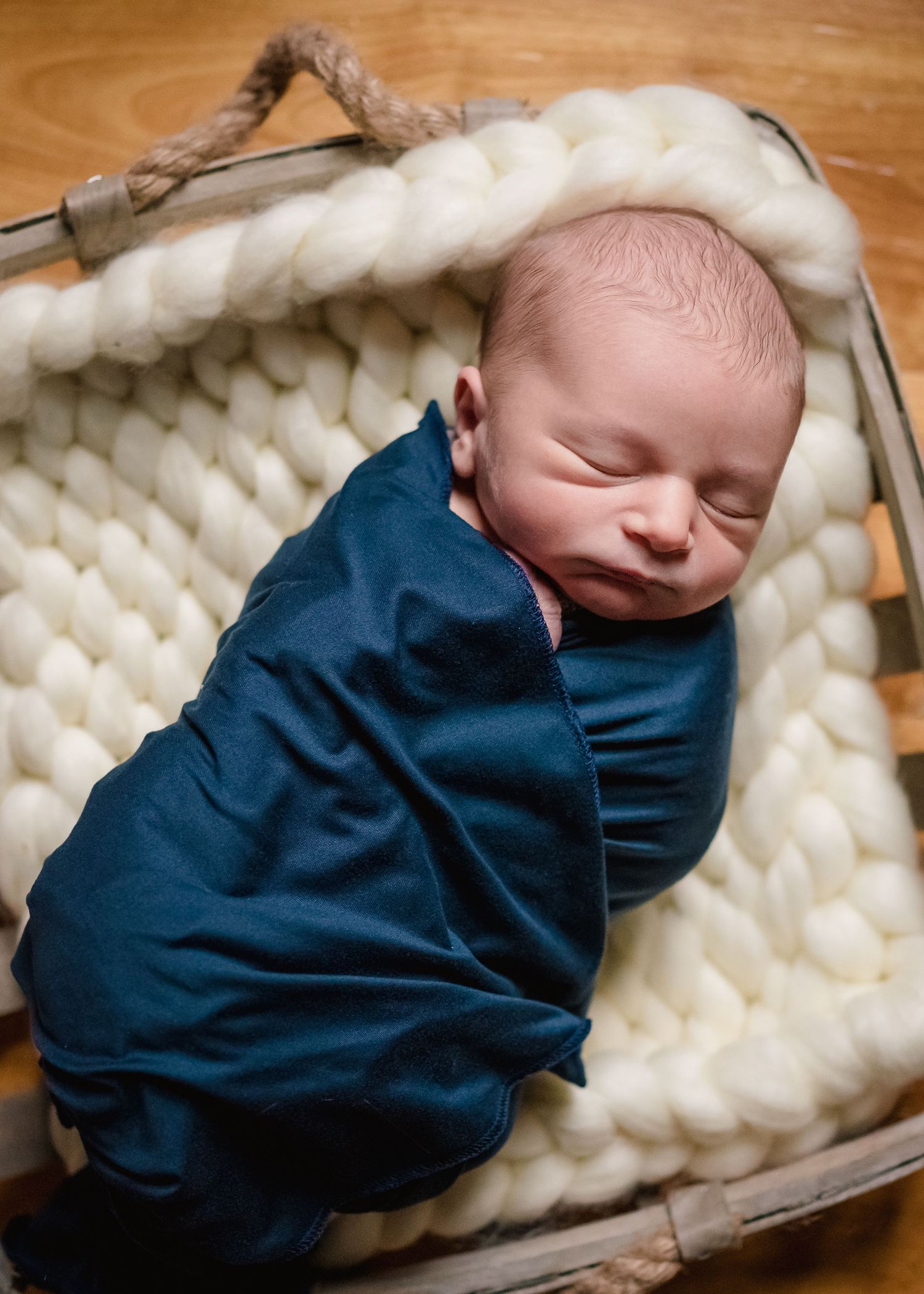 Sleeping newborn wrapped in navy blue swaddle on chunky knit cream blanket.