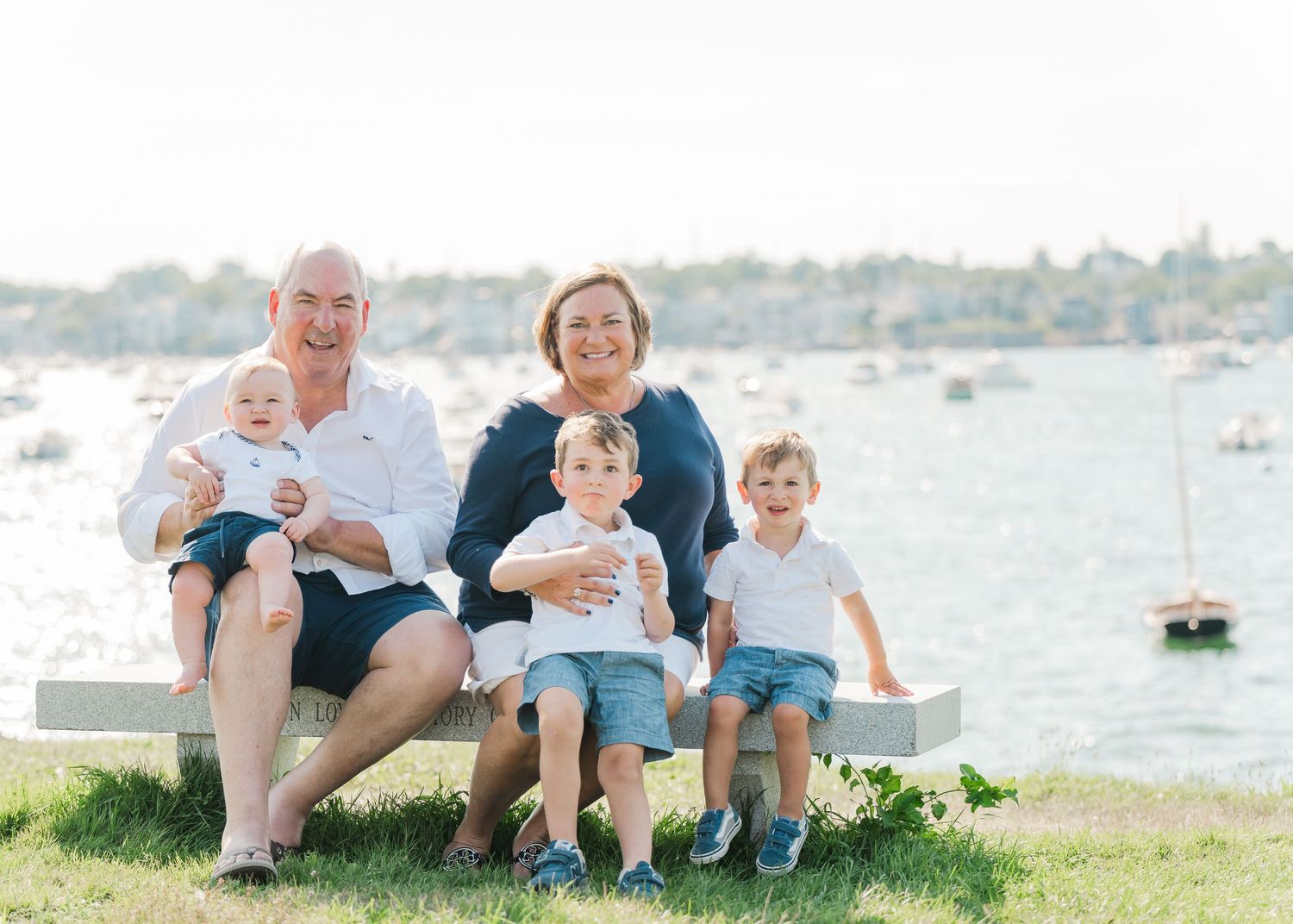 Family relaxing together on grassy shore with boats in harbor on sunny summer day.