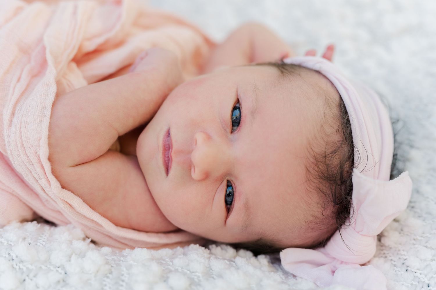 Newborn baby wearing pink headband lying on white textured blanket.