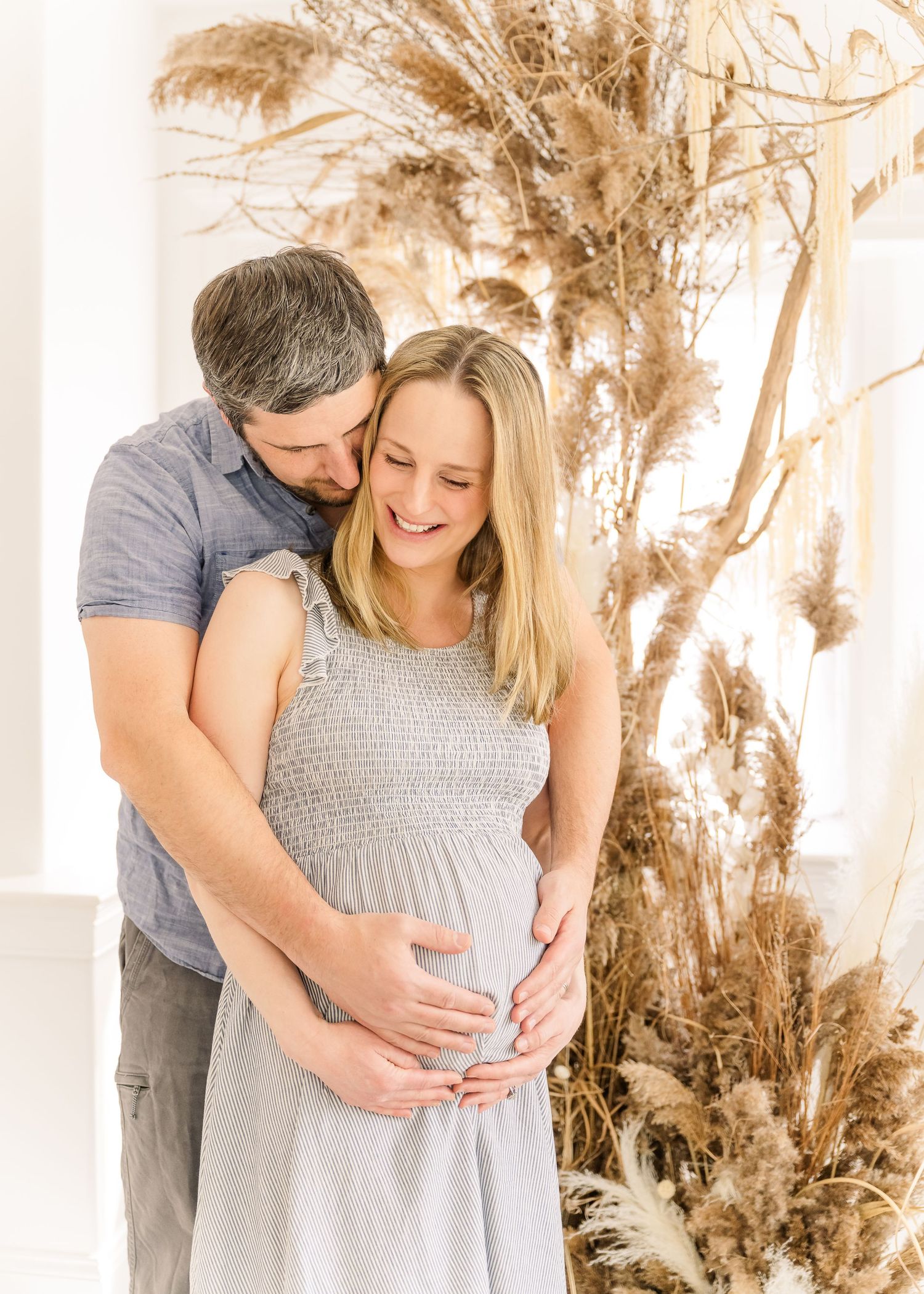 Expecting couple embraces tenderly in a boho setting with dried pampas grass decorations in warm neutral tones.