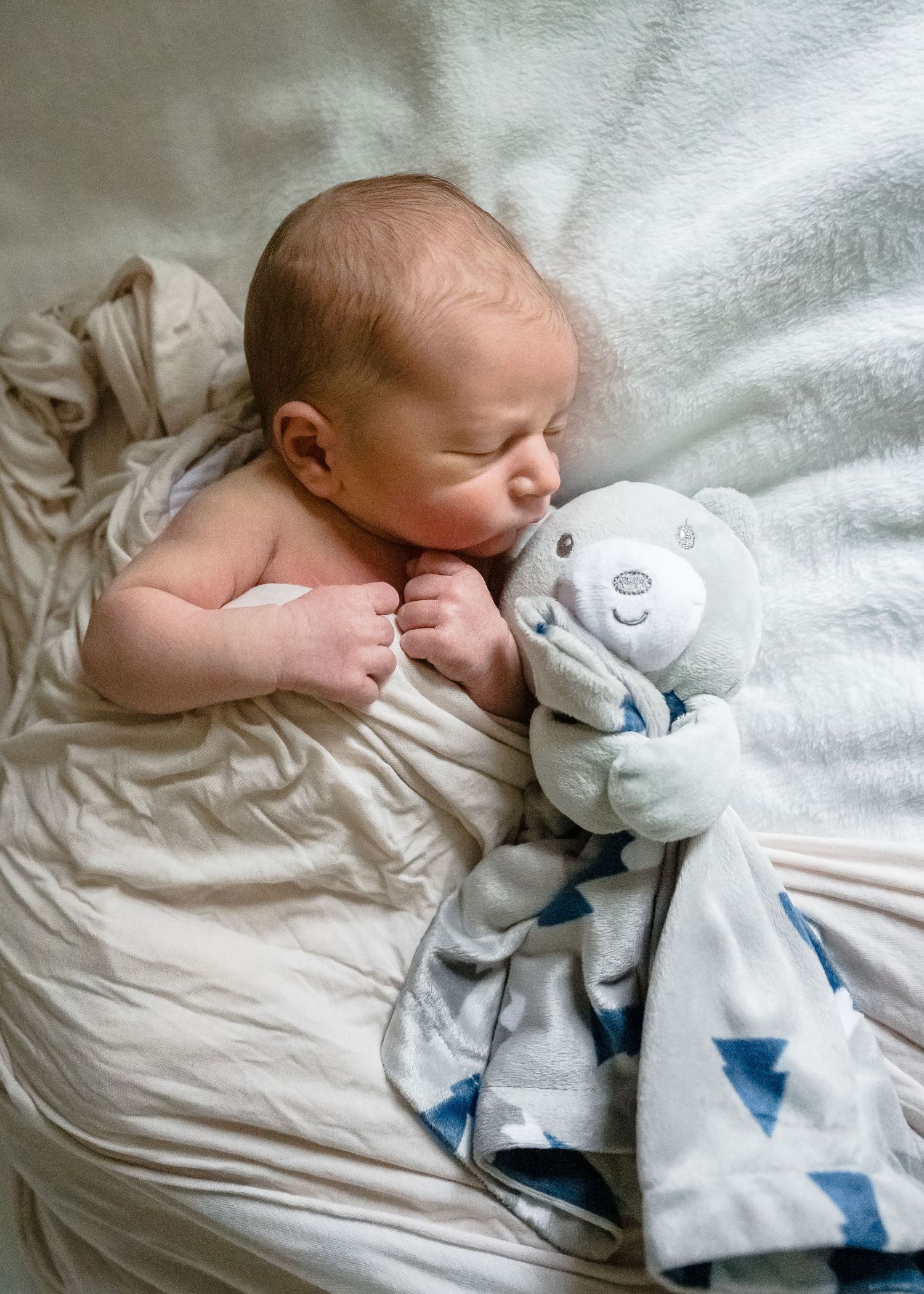 Sleeping baby cuddling with soft plush toy on white bedding.