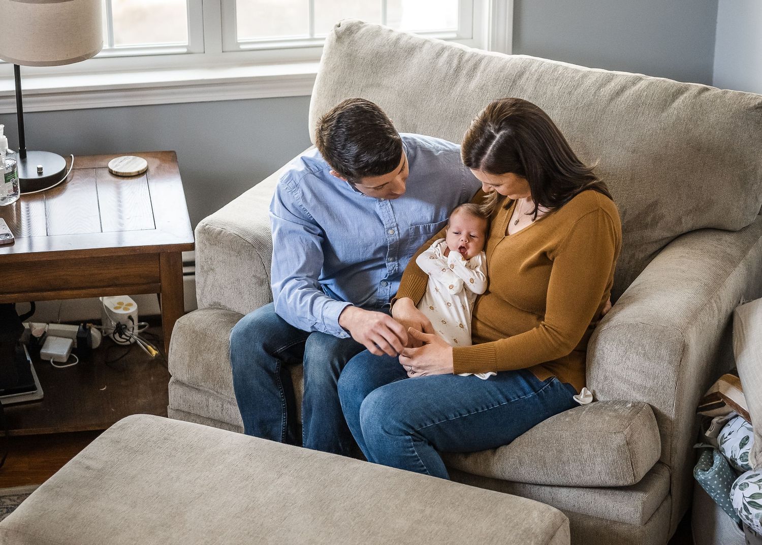Parents cuddle with their newborn baby on a cozy gray sofa in a sunlit living room.