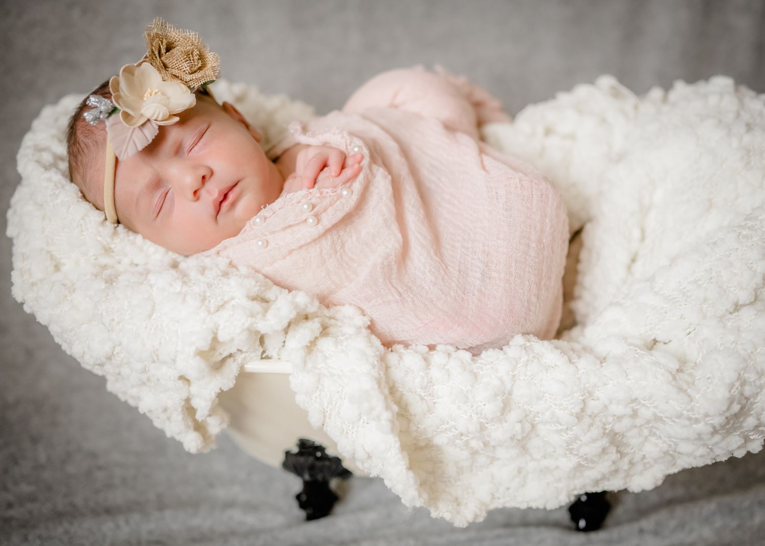 Sleeping newborn in pink wrap with floral headband resting on white textured backdrop.