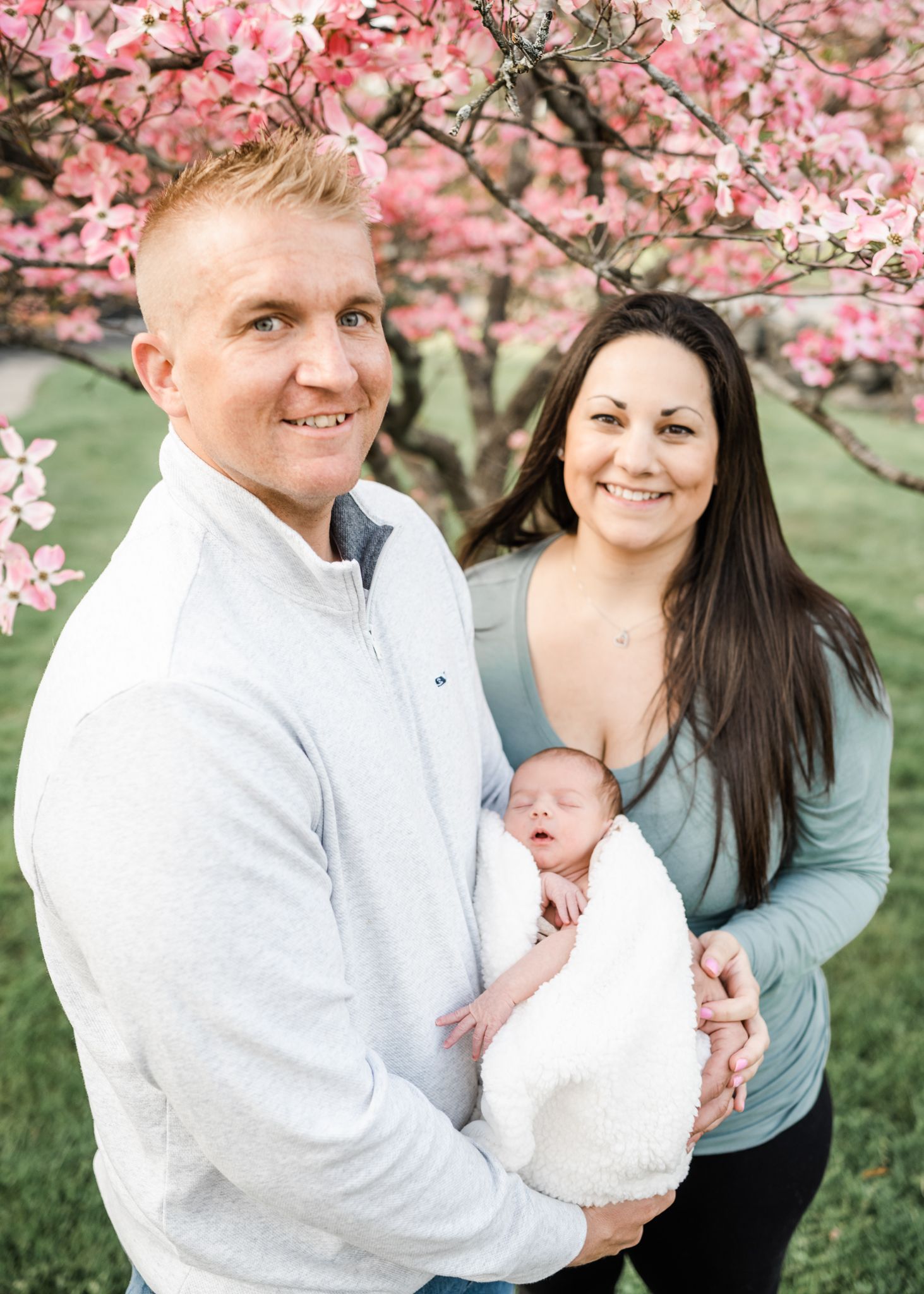A couple holds their newborn baby while standing under blooming pink cherry blossoms in springtime.