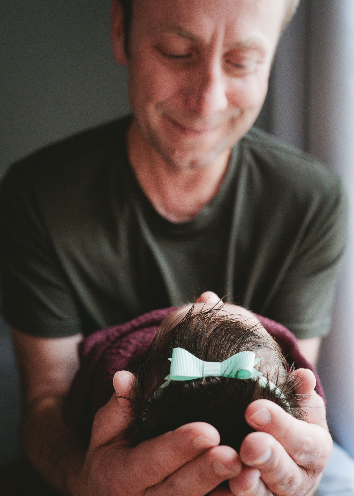 Person in gray shirt holding and gazing lovingly at a swaddled newborn with mint bow.